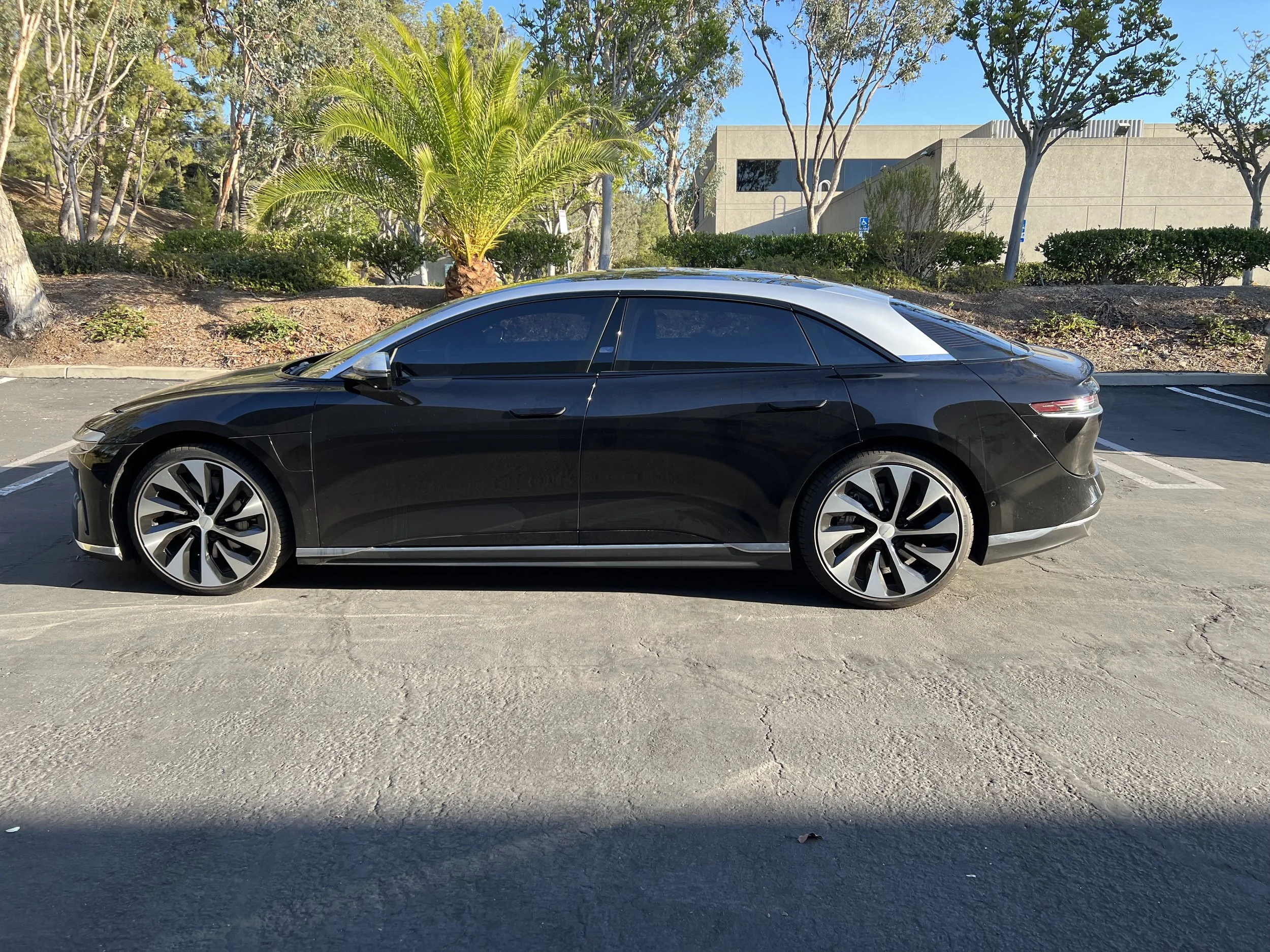 A sleek black electric luxury sedan with tinted windows parked in an outdoor parking lot with trees, bushes, and a building in the background.