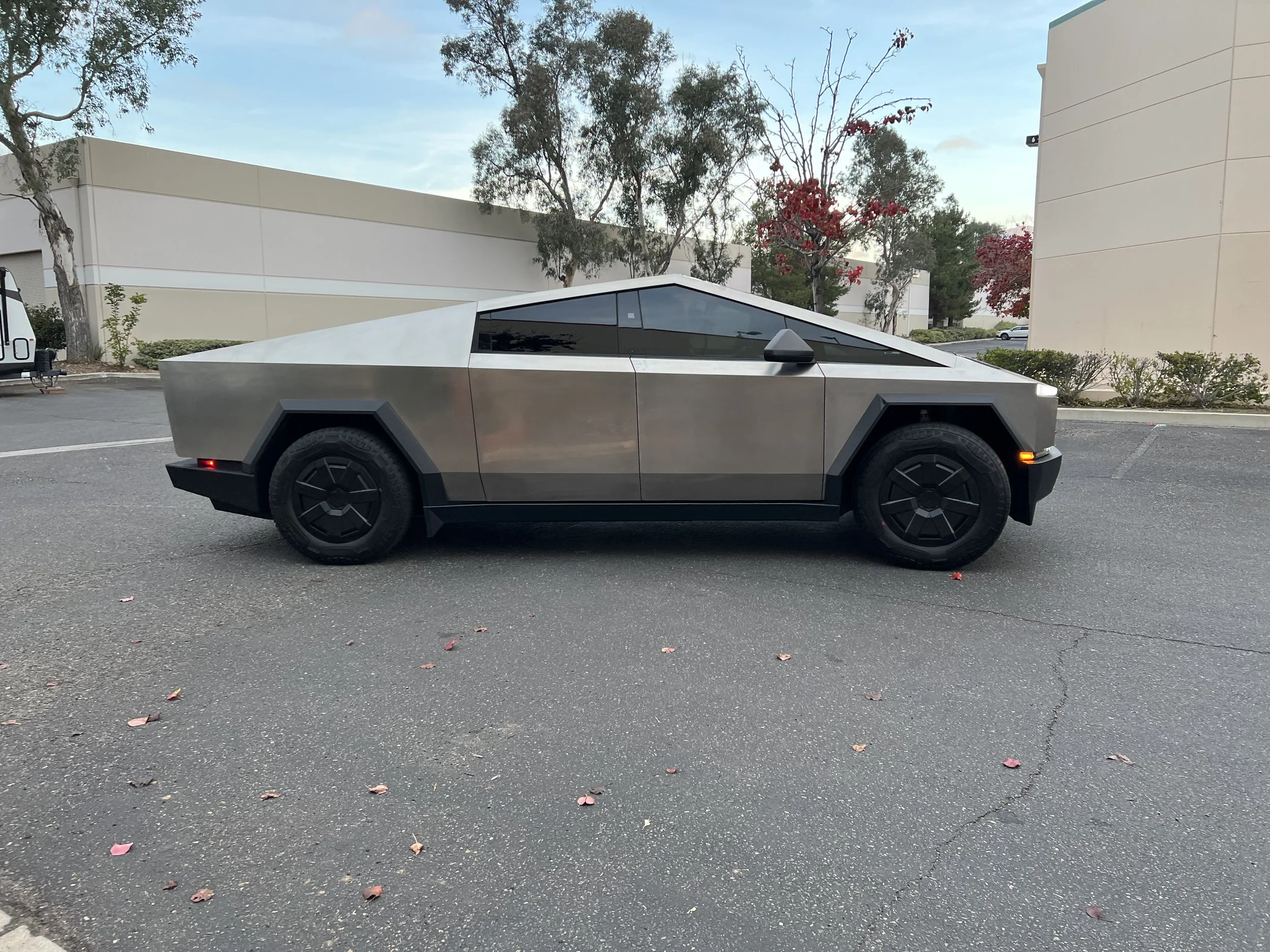 A silver Tesla Cybertruck parked on an asphalt lot outdoors near trees and buildings.