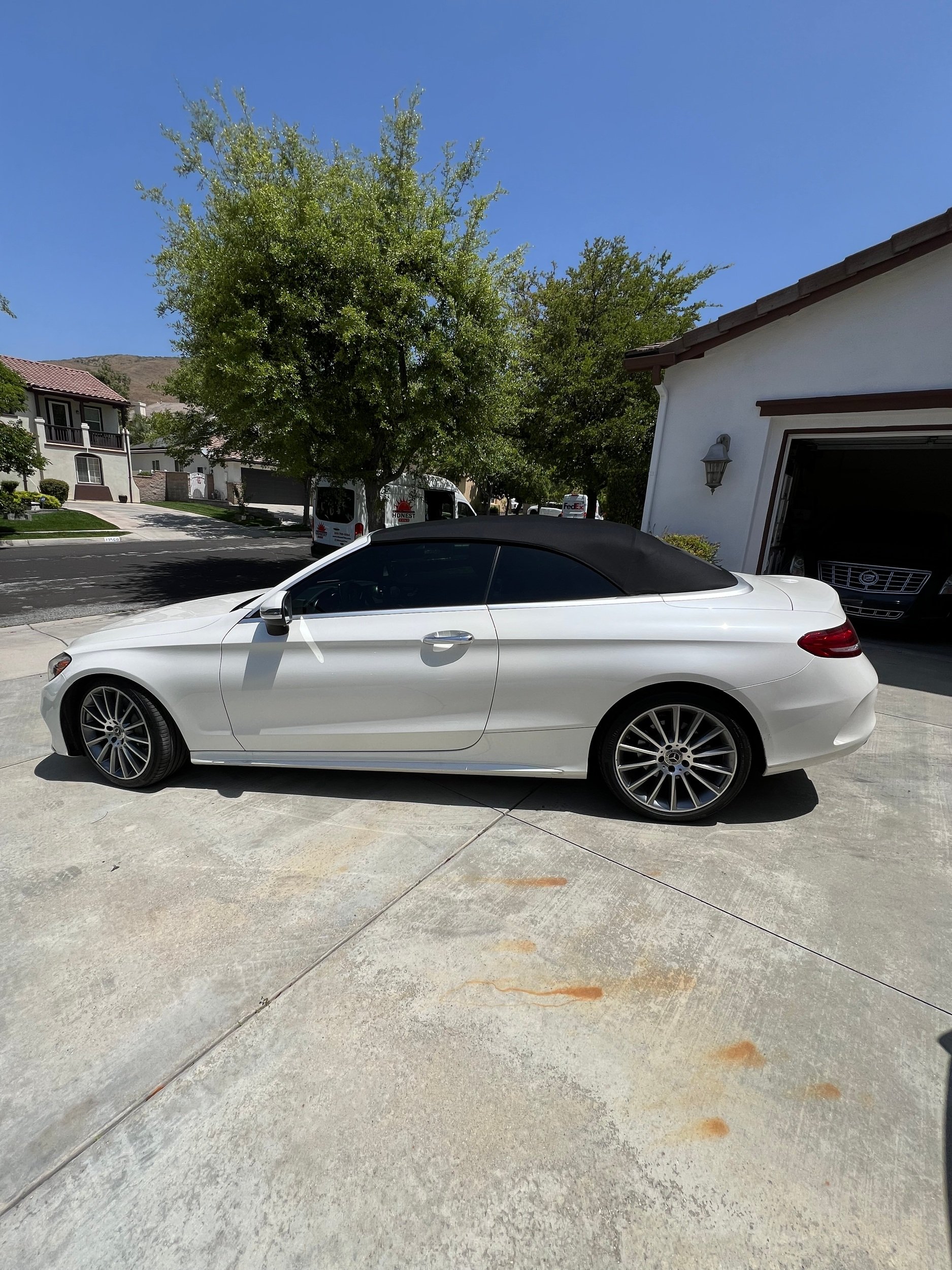 A white convertible car with a black soft top parked in a driveway next to a white garage with open door, trees, and a suburban street background.
