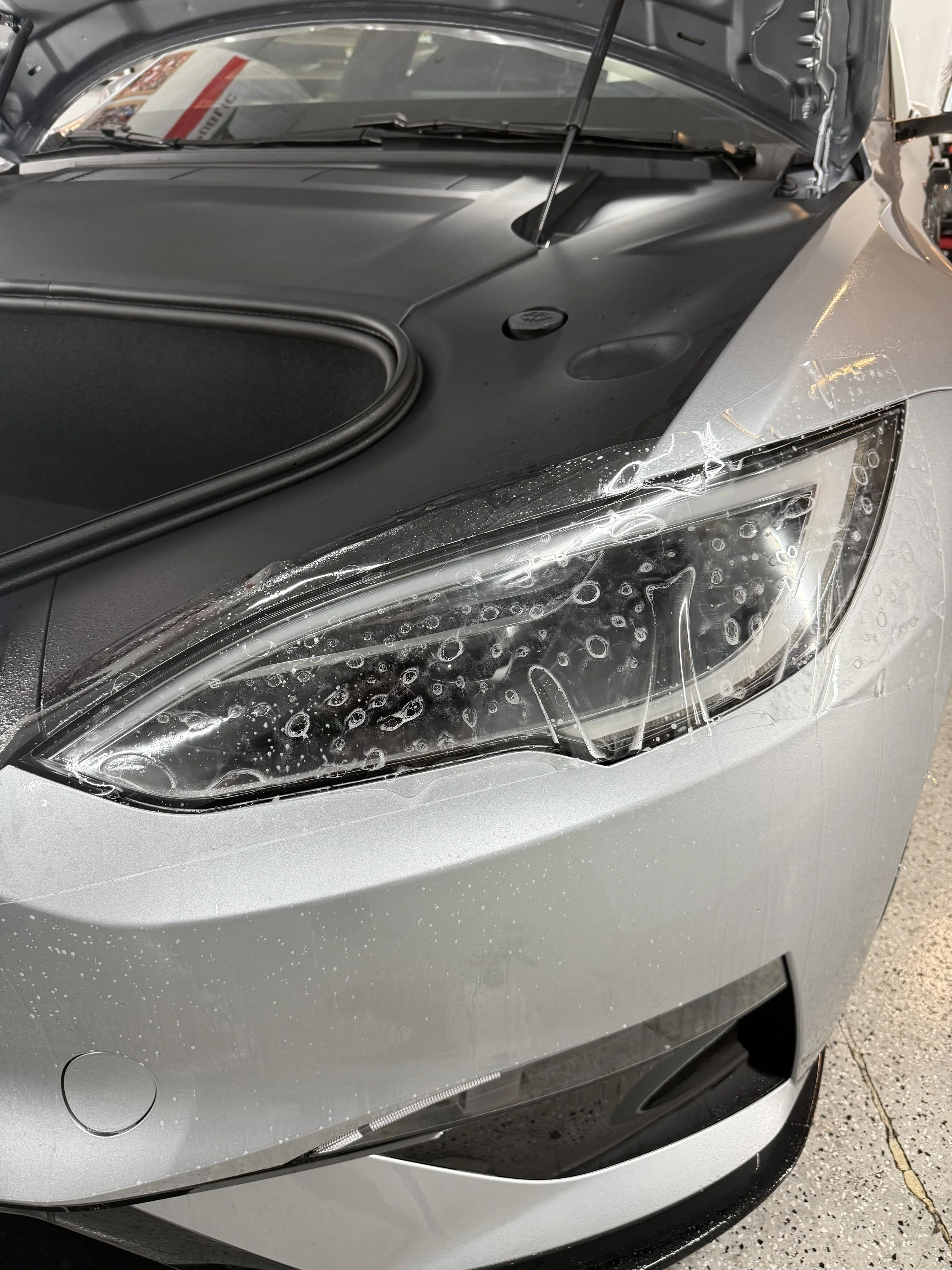 Close-up of the front of a silver car, showing the headlight, part of the hood, and the water droplets on the surface.