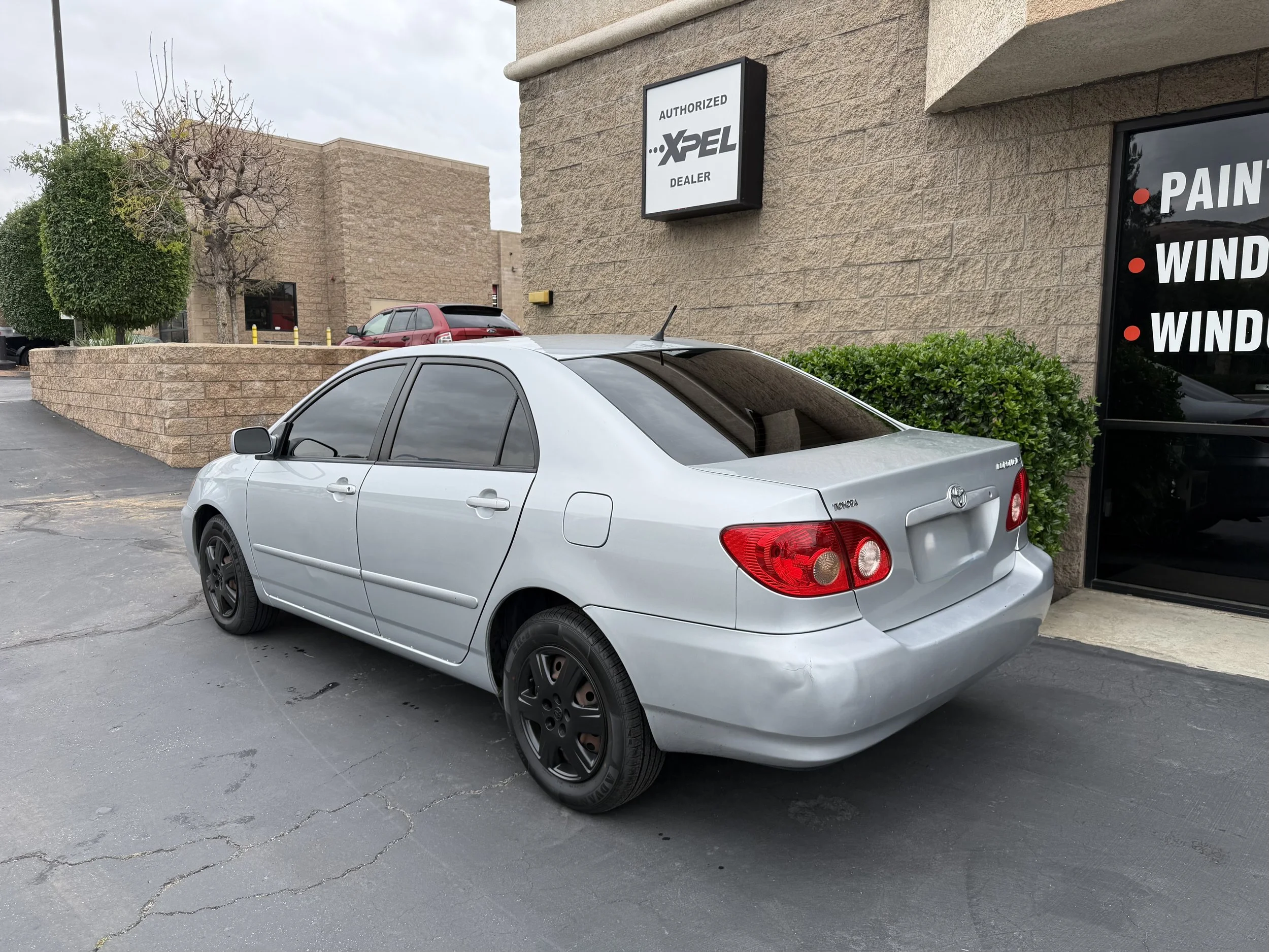 A silver sedan car parked in a parking lot next to a brick wall and bushes, with tinted windows and black wheels.