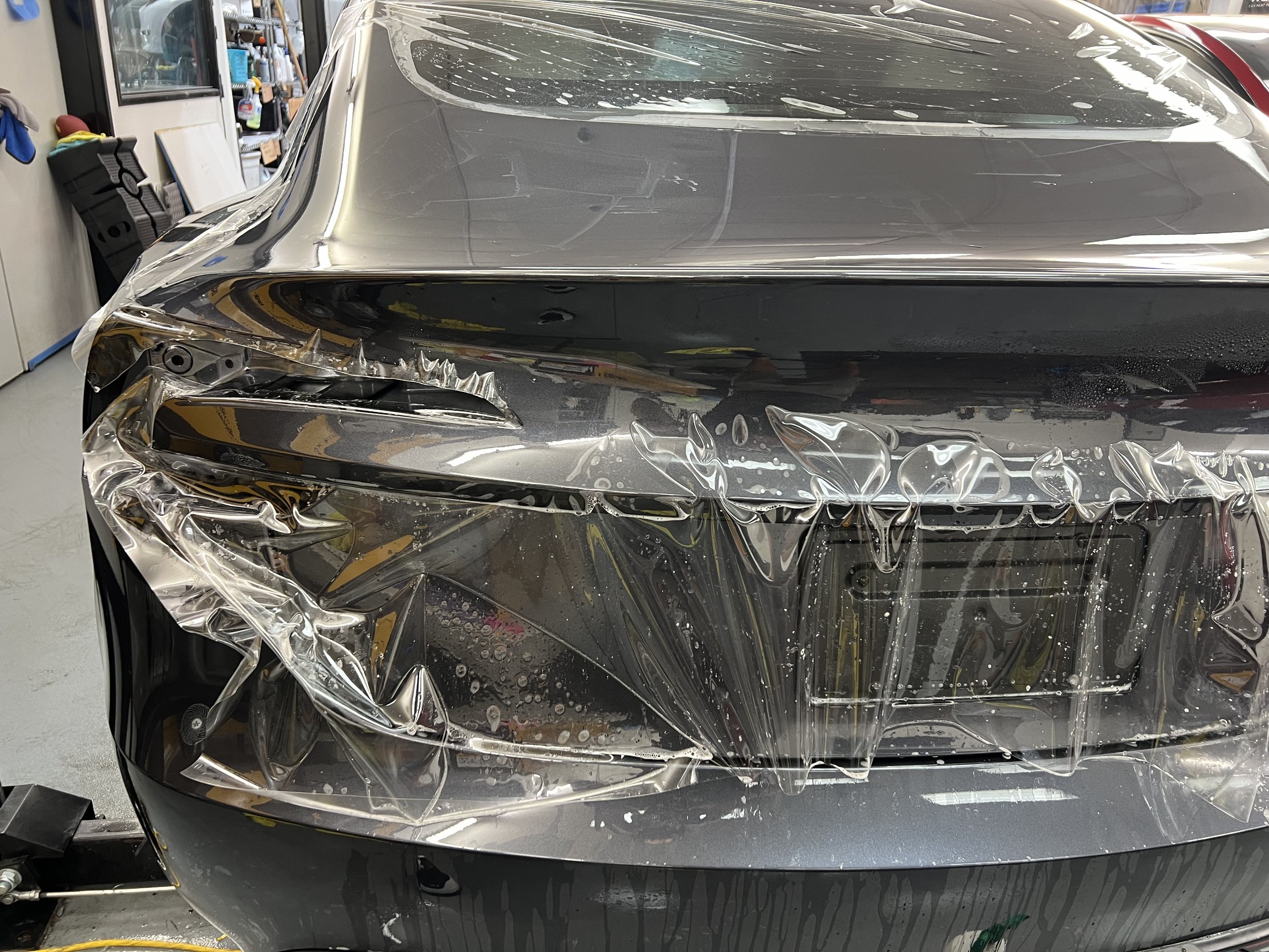 Front of a silver car with damaged headlight and plastic protective covering, in a workshop or garage setting.
