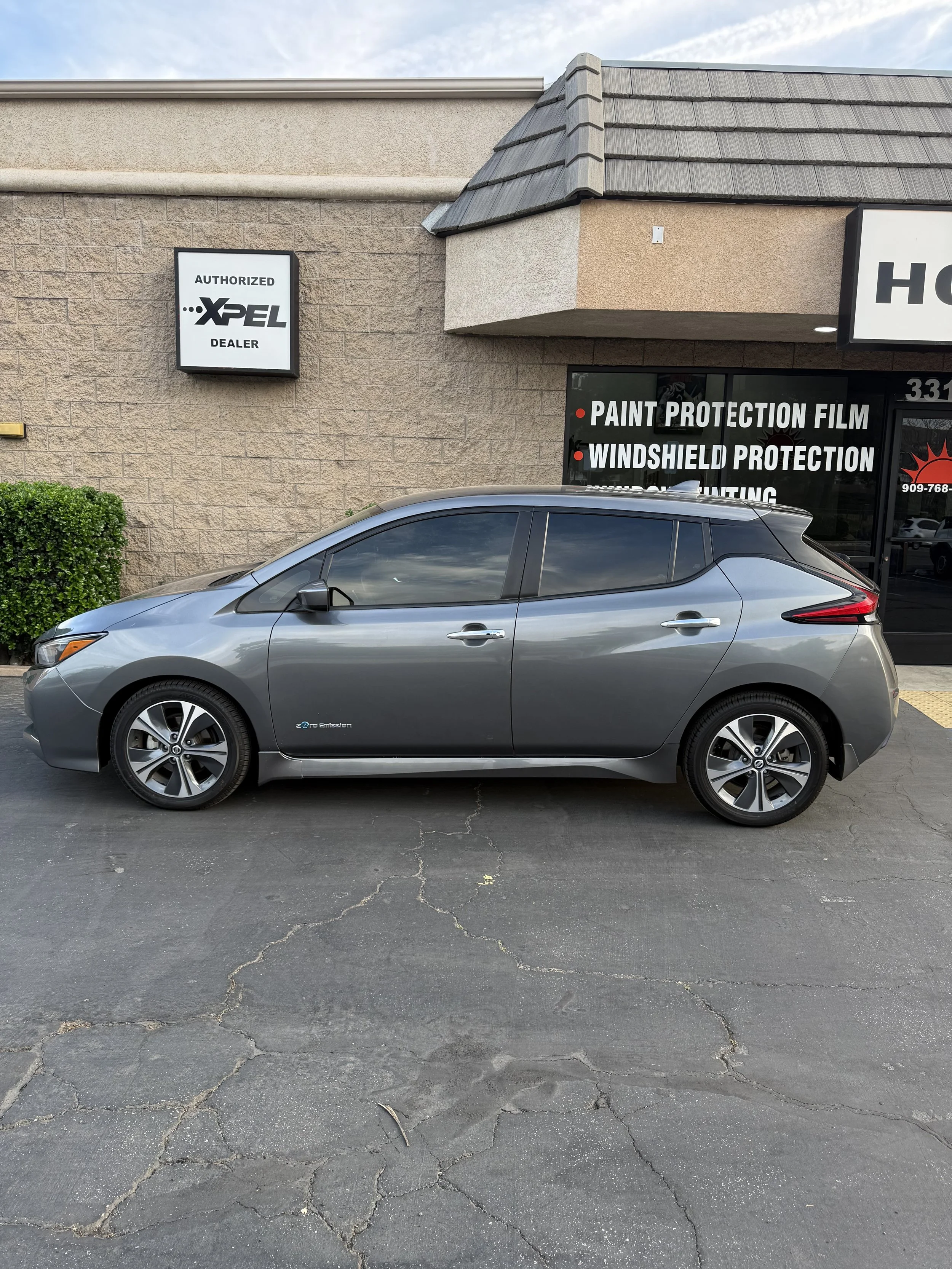 A silver electric car parked outside a dealership with signs for paint protection, windshield protection, and tinting.