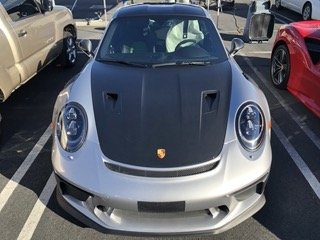 Front view of a silver Porsche sports car with a black hood in a parking lot.