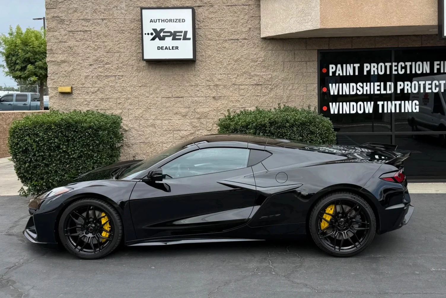 Black sports car with yellow brake calipers parked outside an auto dealership with signs for services such as paint protection film, windshield protection, and window tinting.