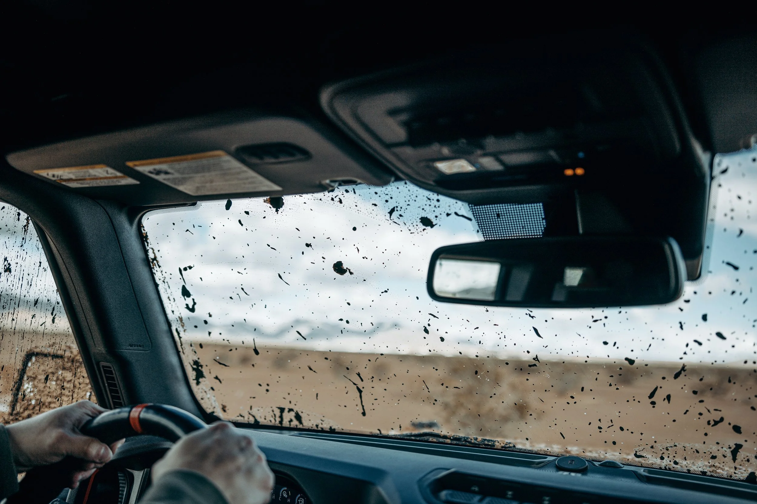 Interior view of a vehicle cockpit with a muddy windshield, showing the steering wheel and rearview mirror, outside a dirt road or field with scattered mud splatters on the windshield.