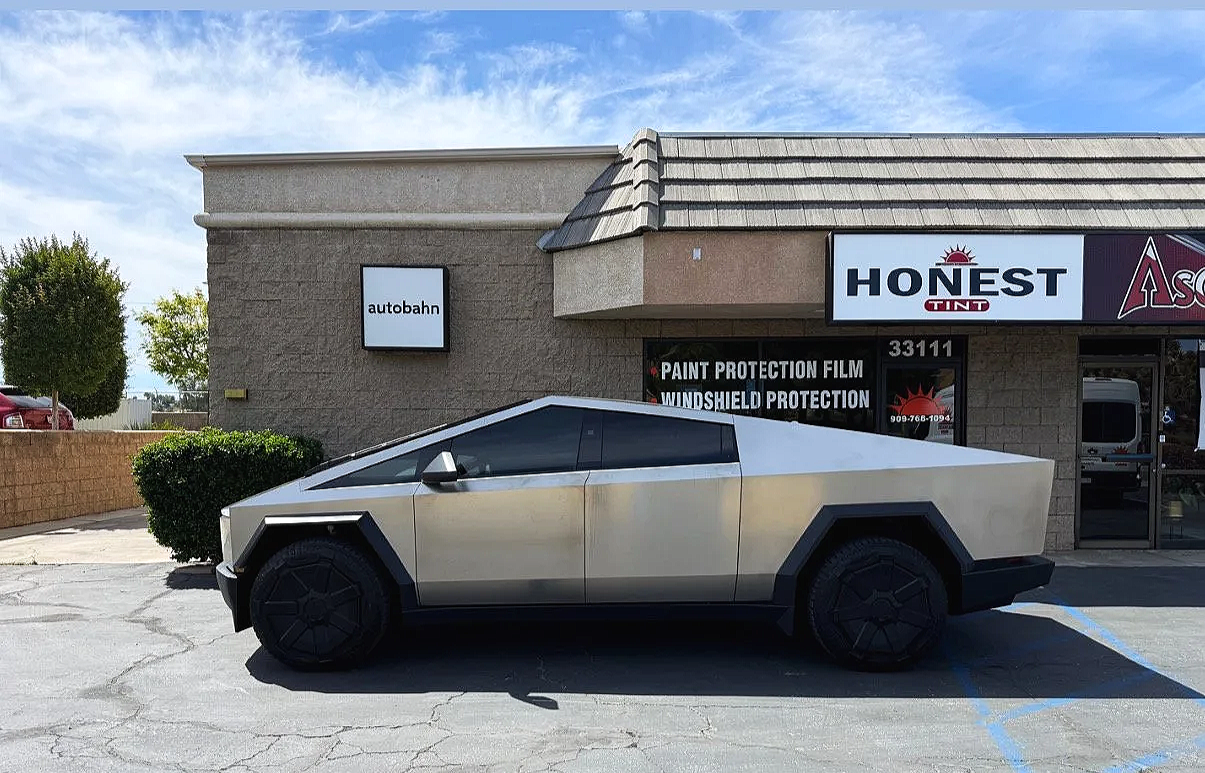 A silver, angular, futuristic-looking car parked in front of a strip mall with signs advertising paint protection, windshield protection, and tint services.