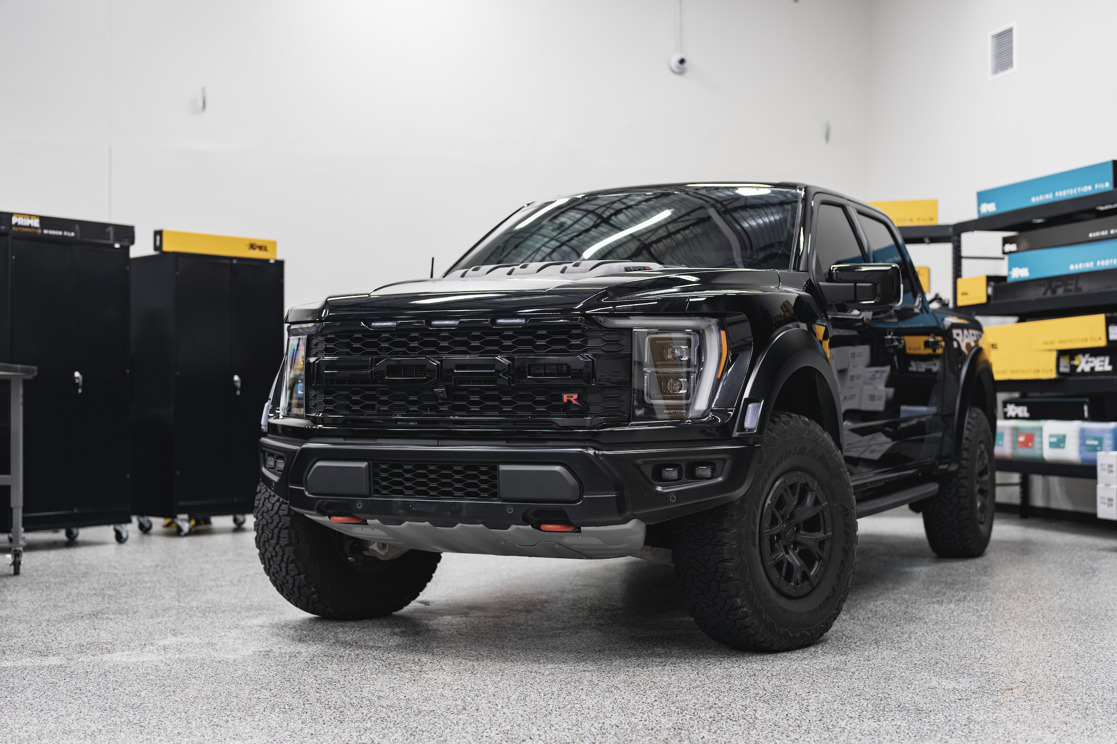 Black Ford Raptor pickup truck in an indoor storage or showroom area with shelves of automotive supplies and storage cabinets.