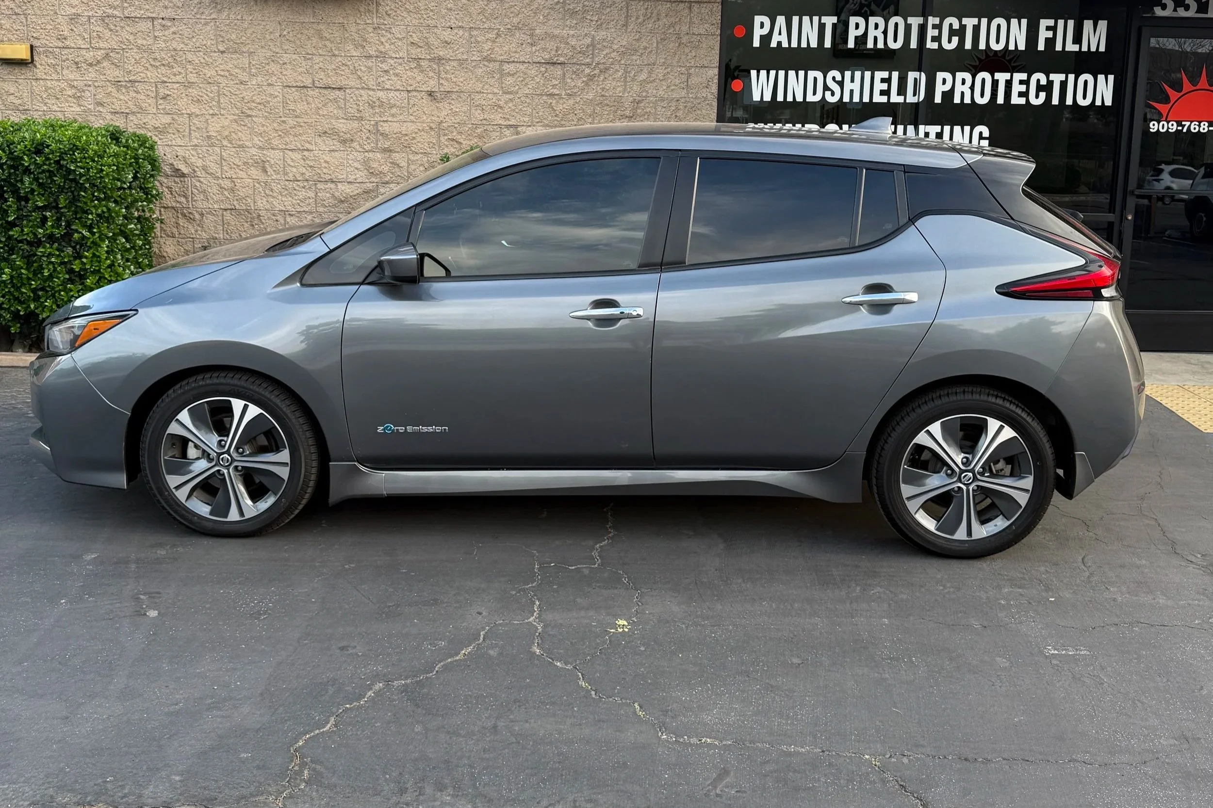 A silver electric hatchback car parked in front of a building with a sign listing services including paint protection film, windshield protection, and window tinting.
