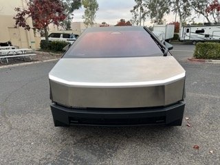 Front view of a silver vintage car parked in a lot with trees and buildings in the background.