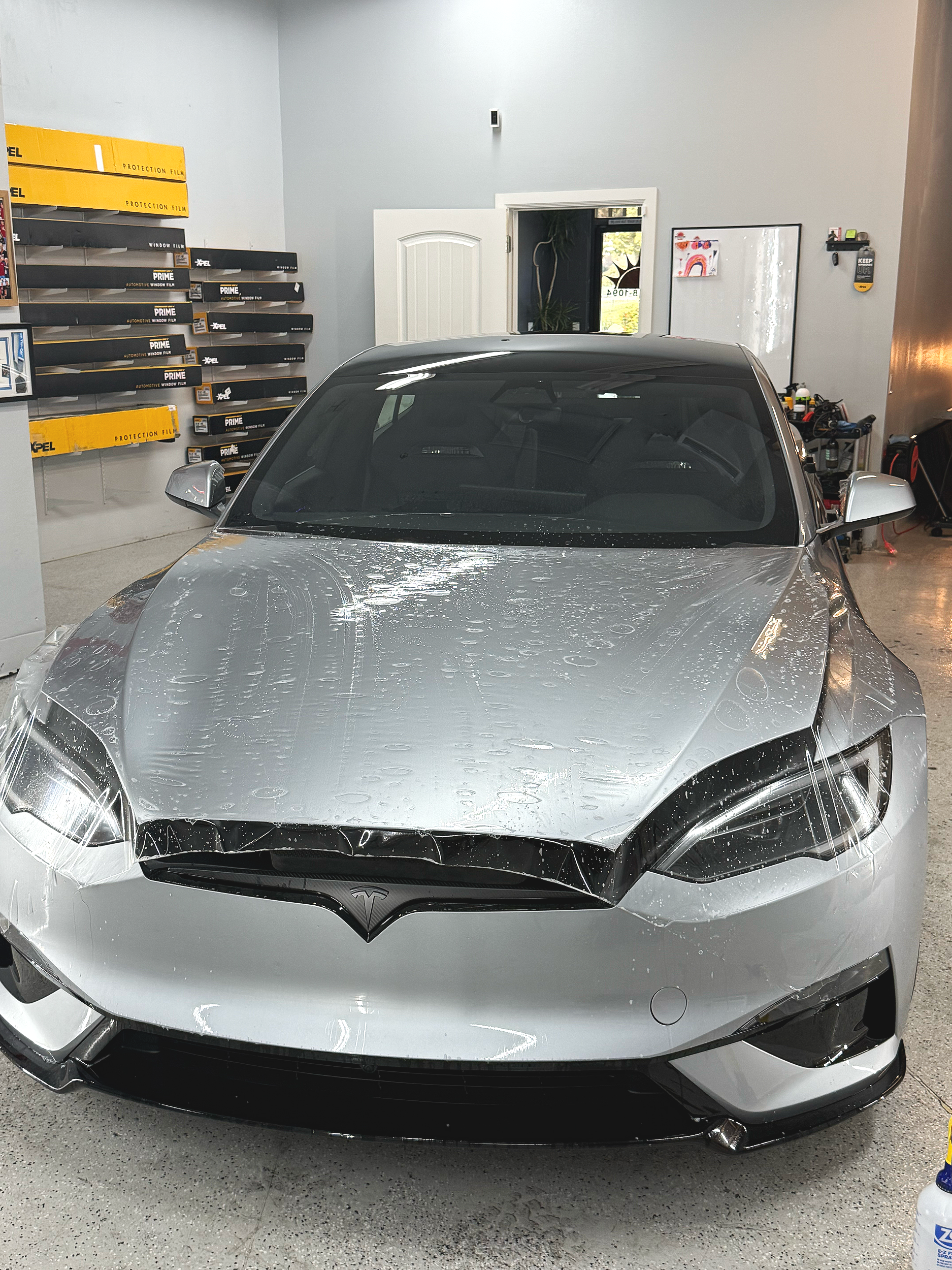 A silver Tesla car inside a garage with water droplets on the hood. The garage has wall-mounted shelves with car film and protection film in yellow and black boxes. There is a small window, a white door, and various tools and bottles on a shelf to the right.