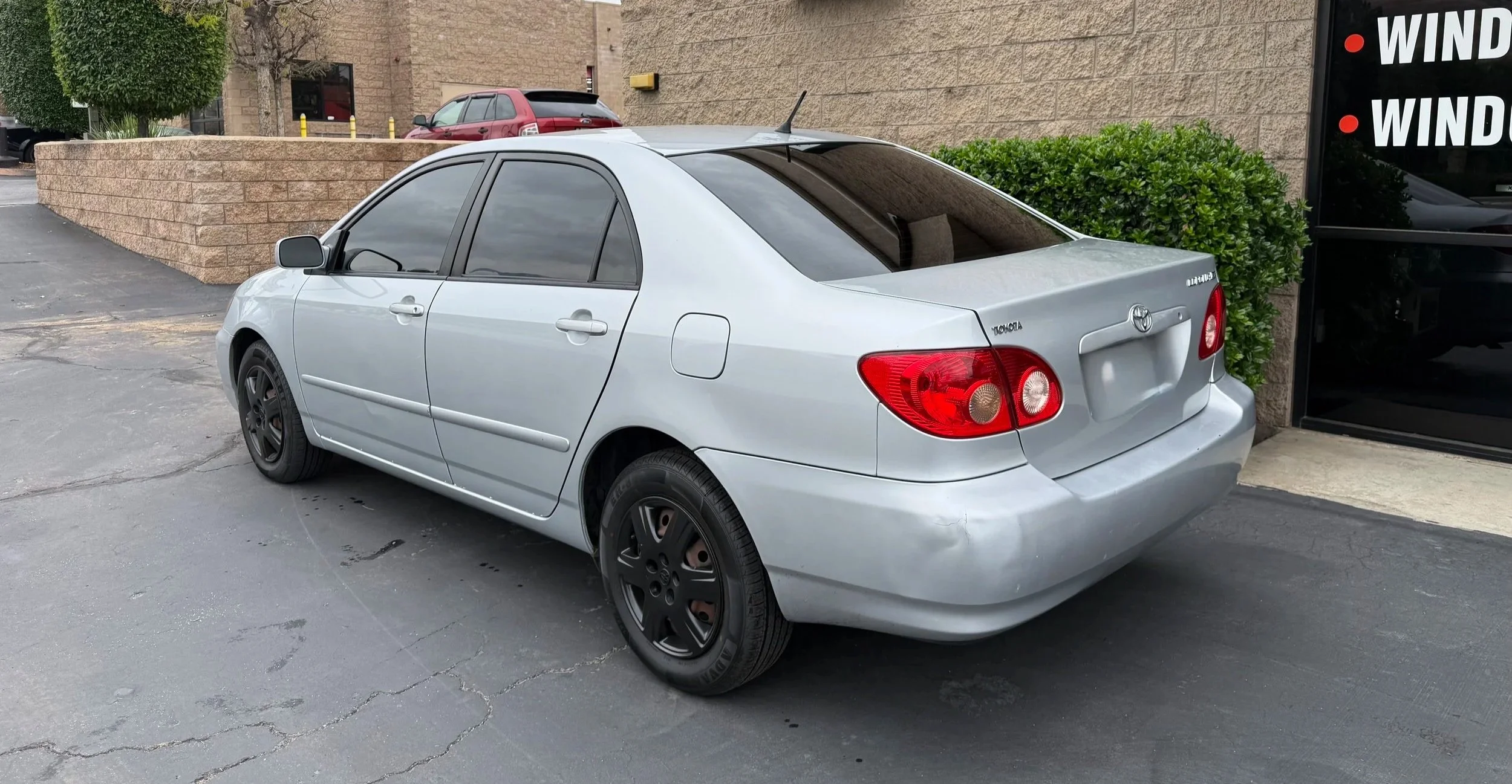 Silver compact sedan car parked near a building with a brick wall and bushes.