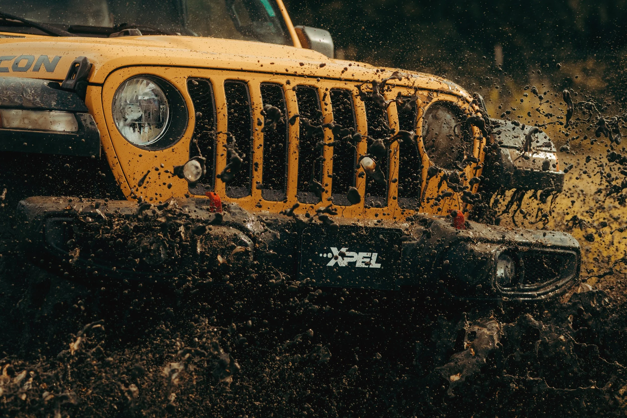 A yellow Jeep driving through muddy terrain, splashing mud around.