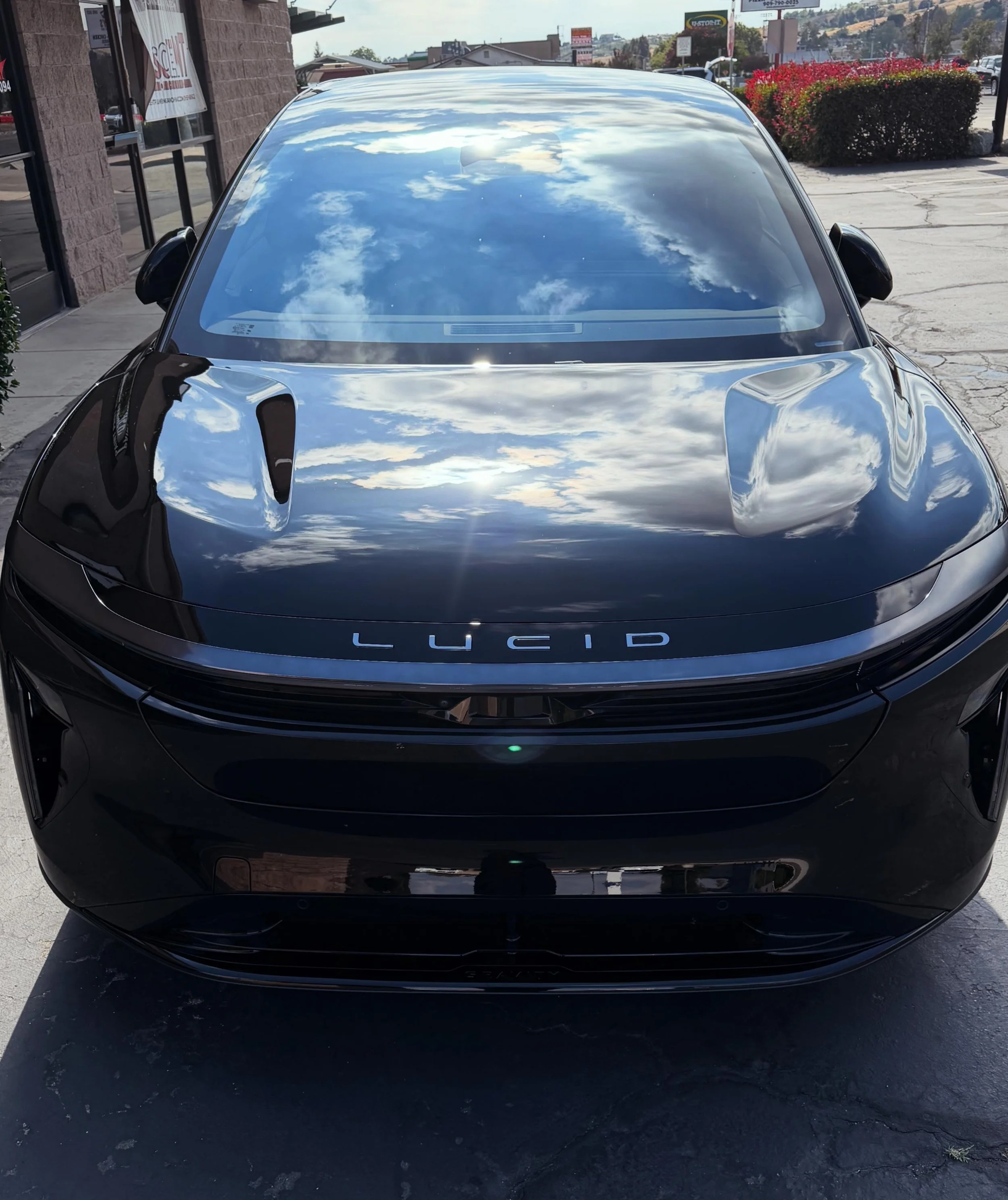 Front view of a black Lucid electric vehicle parked outdoors, with a reflective windshield showing clouds, buildings, and blue sky, and a visible Lucid logo on the front bumper.