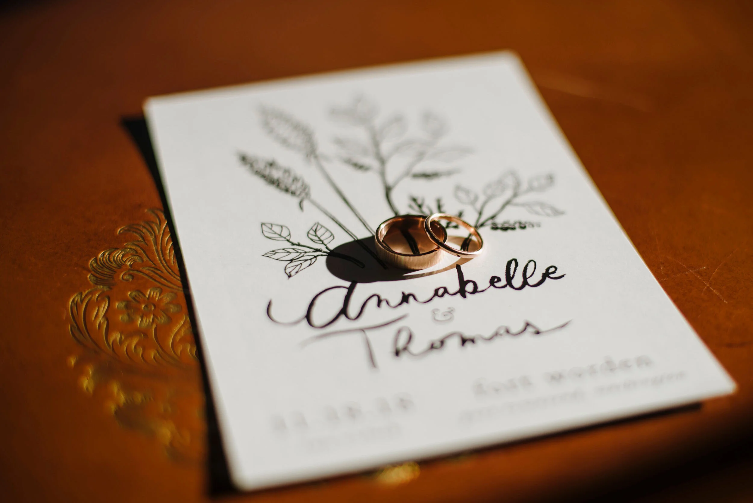 A wedding invitation with black floral line art and the names Anabelle and Thomas handwritten; on top of the invitation, there are two gold wedding rings placed, casting shadows on the paper.