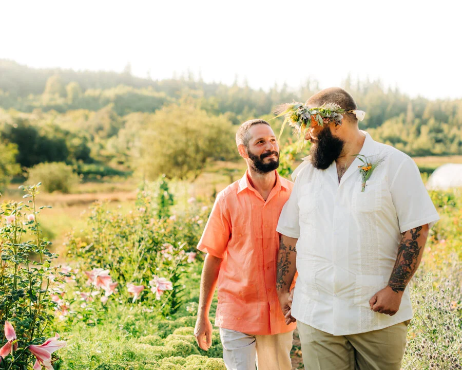 Two men on their wedding day, one wearing a white shirt with tattoos and a flower crown, the other in an orange shirt, with trees and flowers in the background.