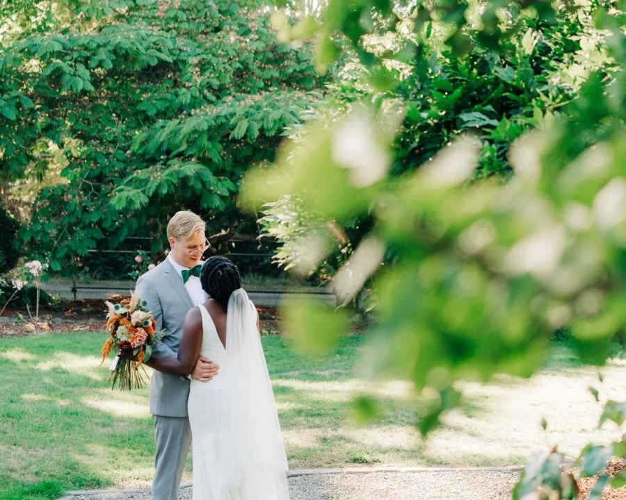 A newlywed couple, the groom in a light gray suit and bow tie, and the bride in a white wedding dress with a veil, standing outdoors on a lawn surrounded by green trees, sharing a tender moment with the bride holding a bouquet of flowers.