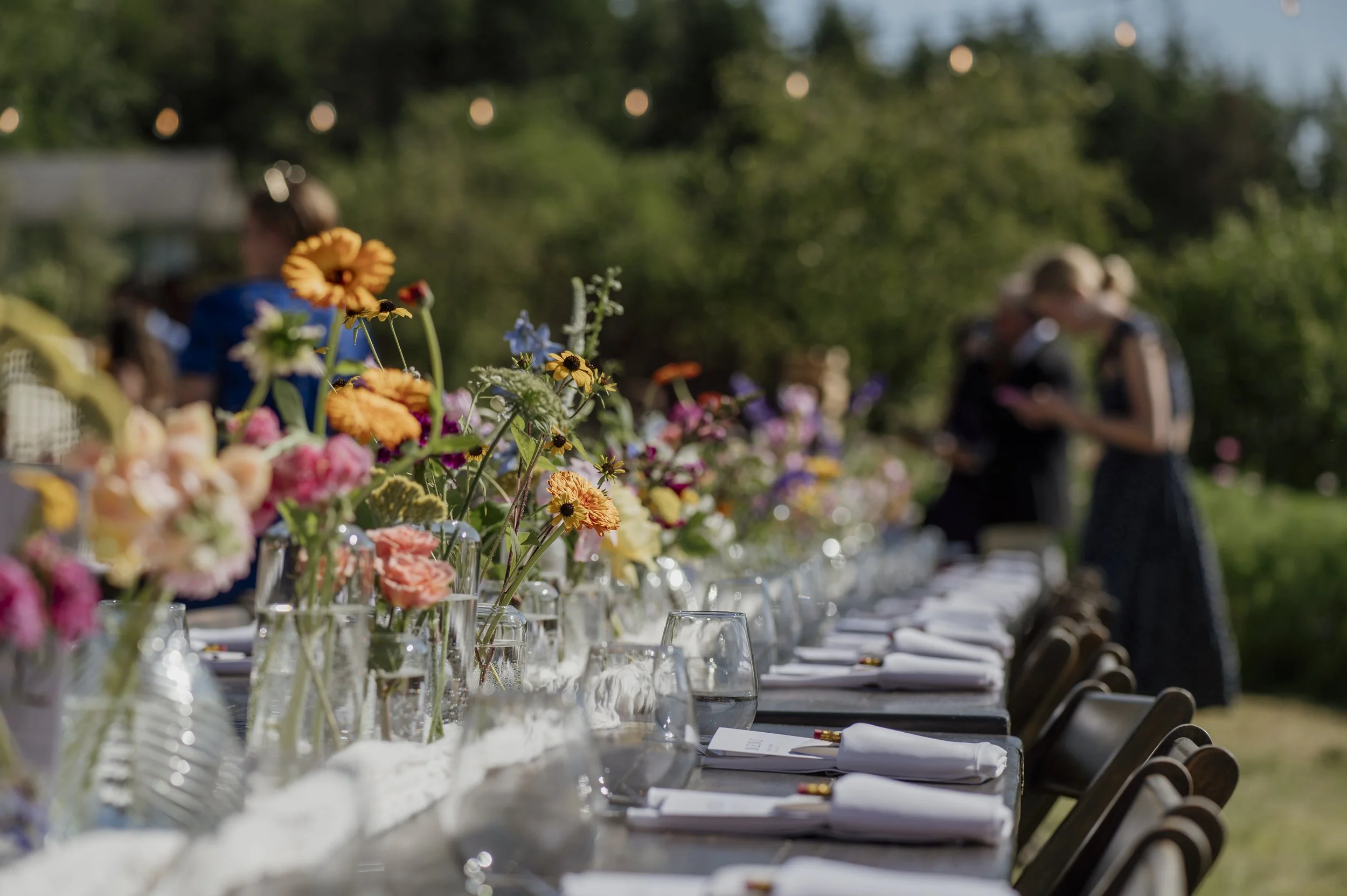 Long table set for outdoor wedding reception with colorful flower arrangements, glasses, and napkins, with people in the background under a string light, during daytime.