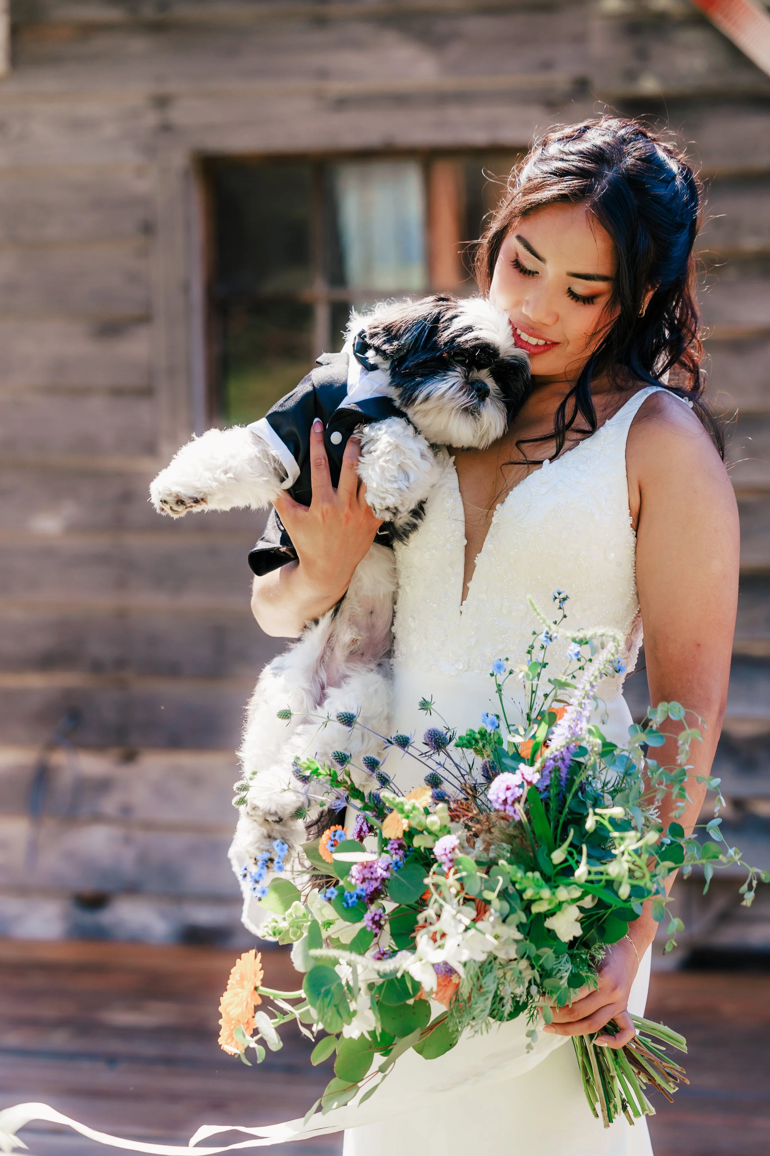 A woman in a wedding dress holding a bouquet of flowers and a small dog dressed in a tuxedo, near a wooden building
