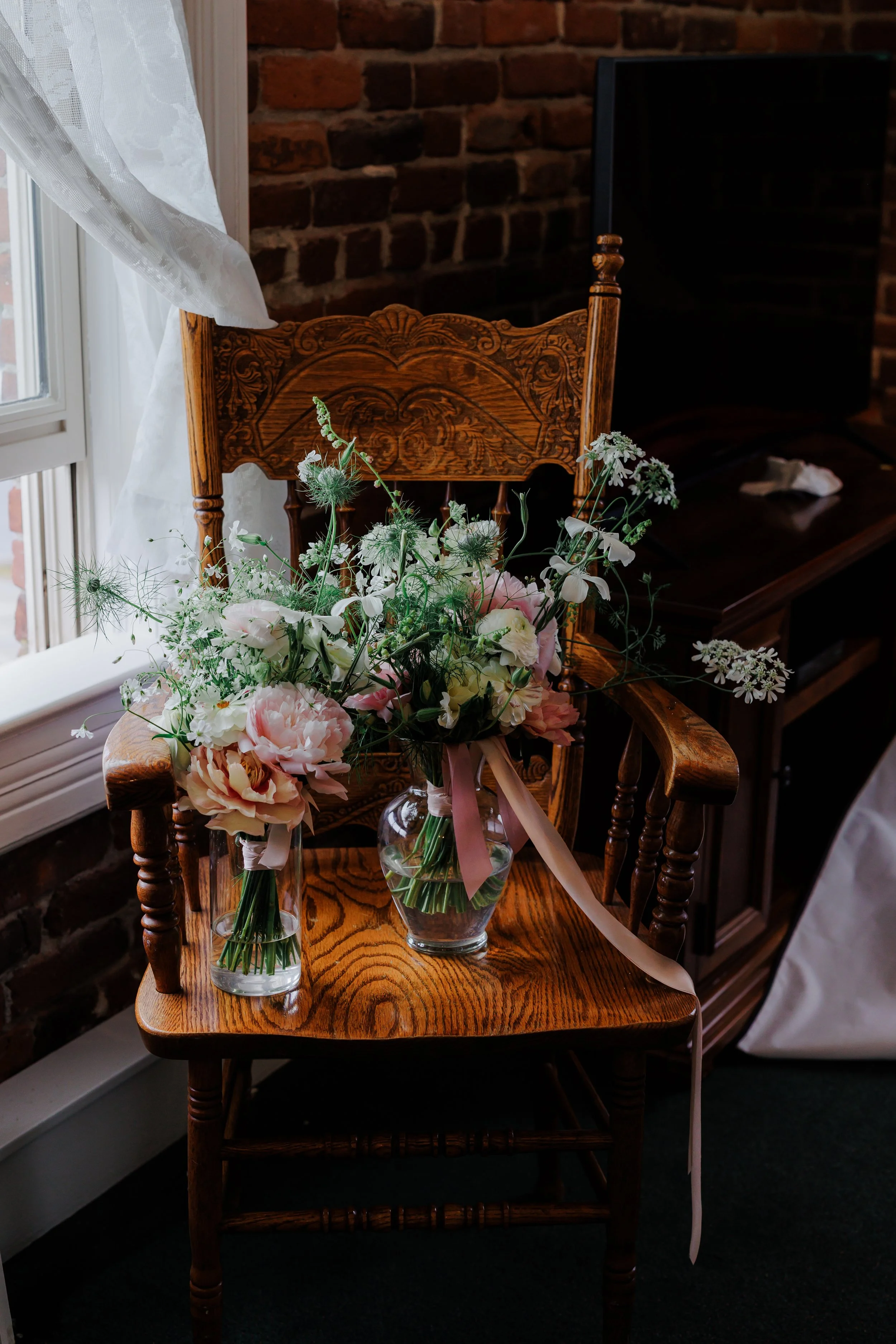 Two glass vases with pink and white flowers on a wooden chair near a window with white curtains, against a brick wall.