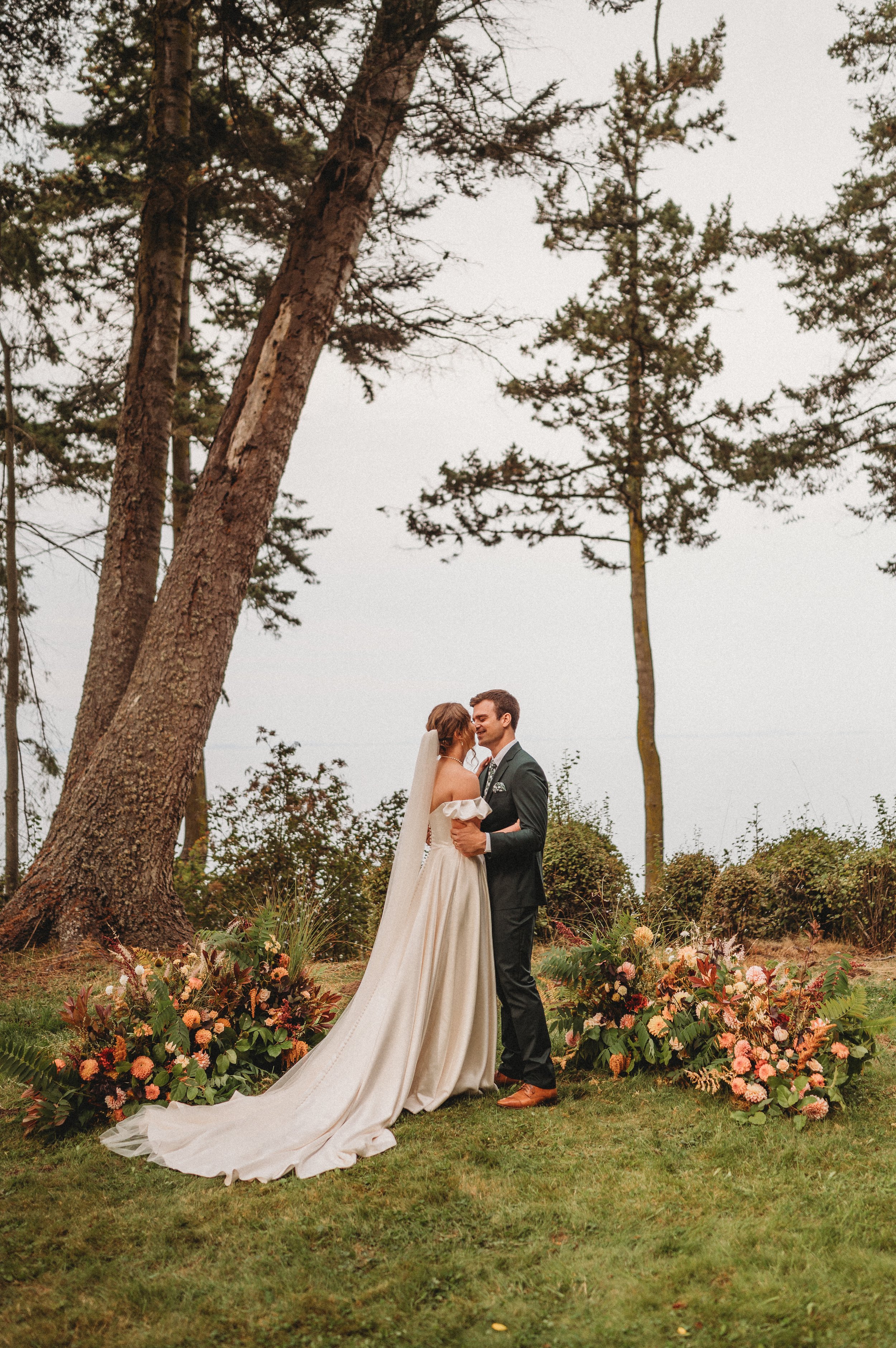 A bride and groom stand together outdoors under tall trees, celebrating their wedding, with floral arrangements on the grass.