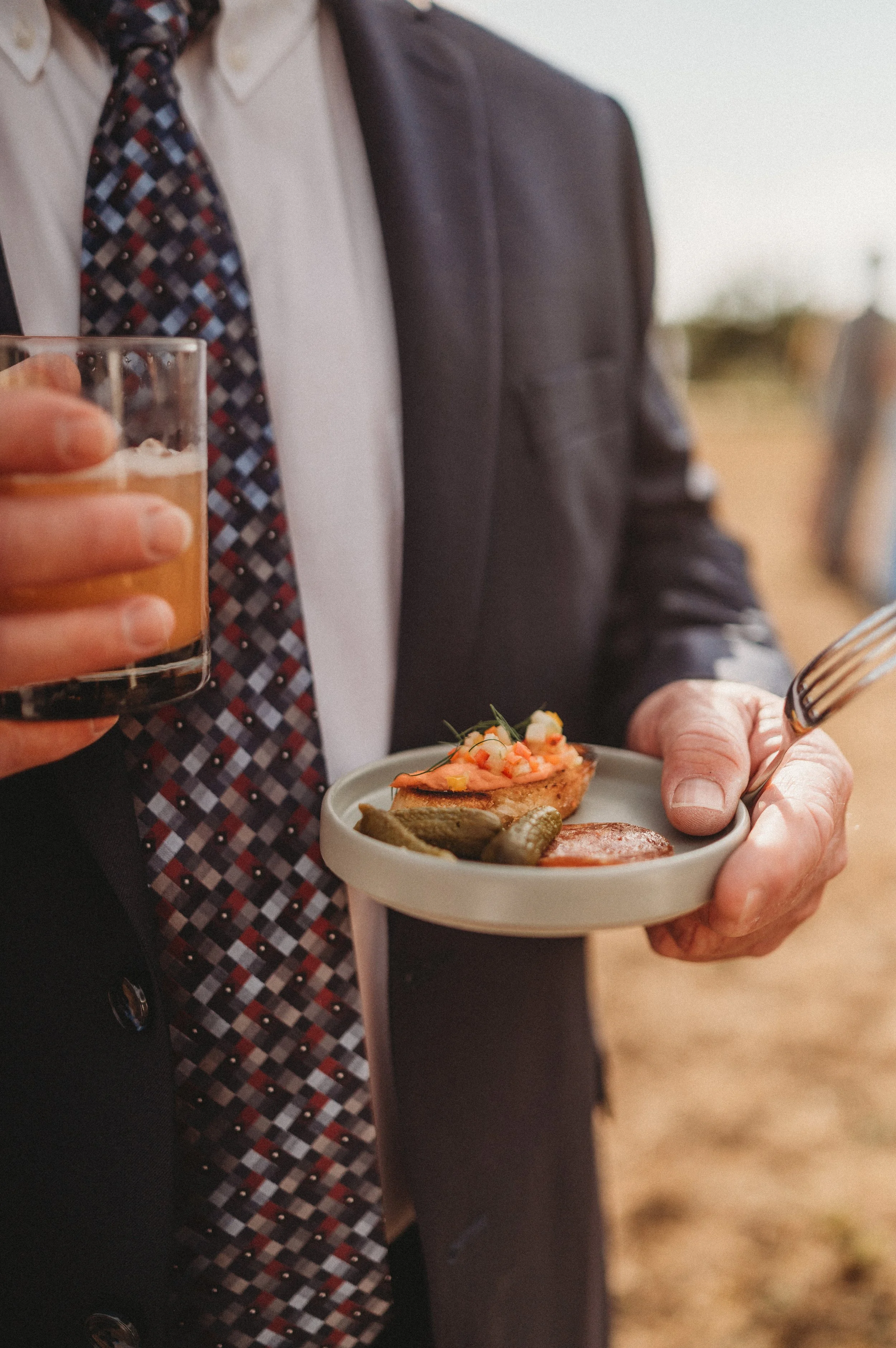 A man in a suit holding a drink and a small plate with snacks, including a piece of toasted bread with toppings, pickles, and a slice of cured meat, at an outdoor event.