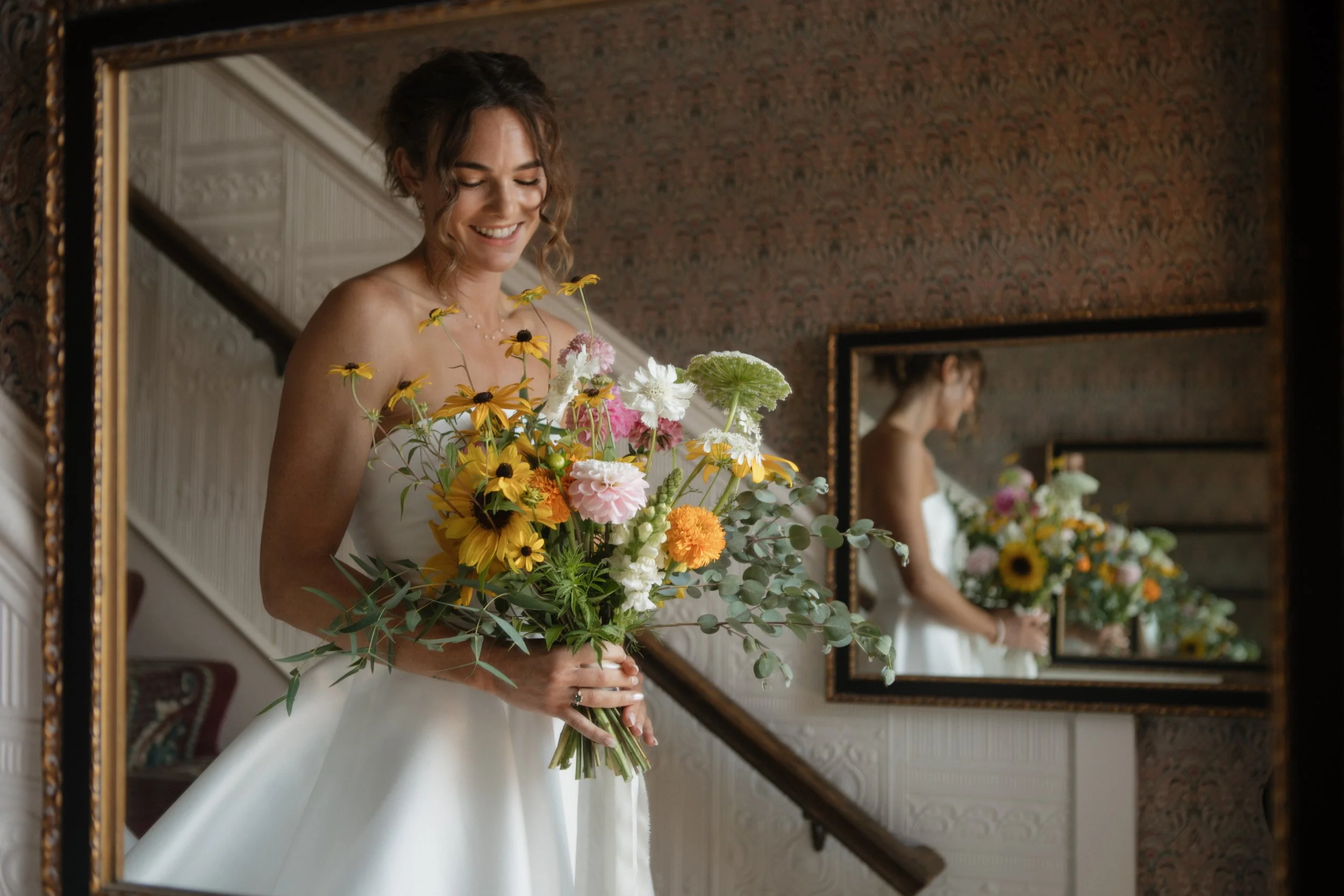 Bride in a white wedding dress holding a colorful bouquet of flowers, reflected in a mirror.