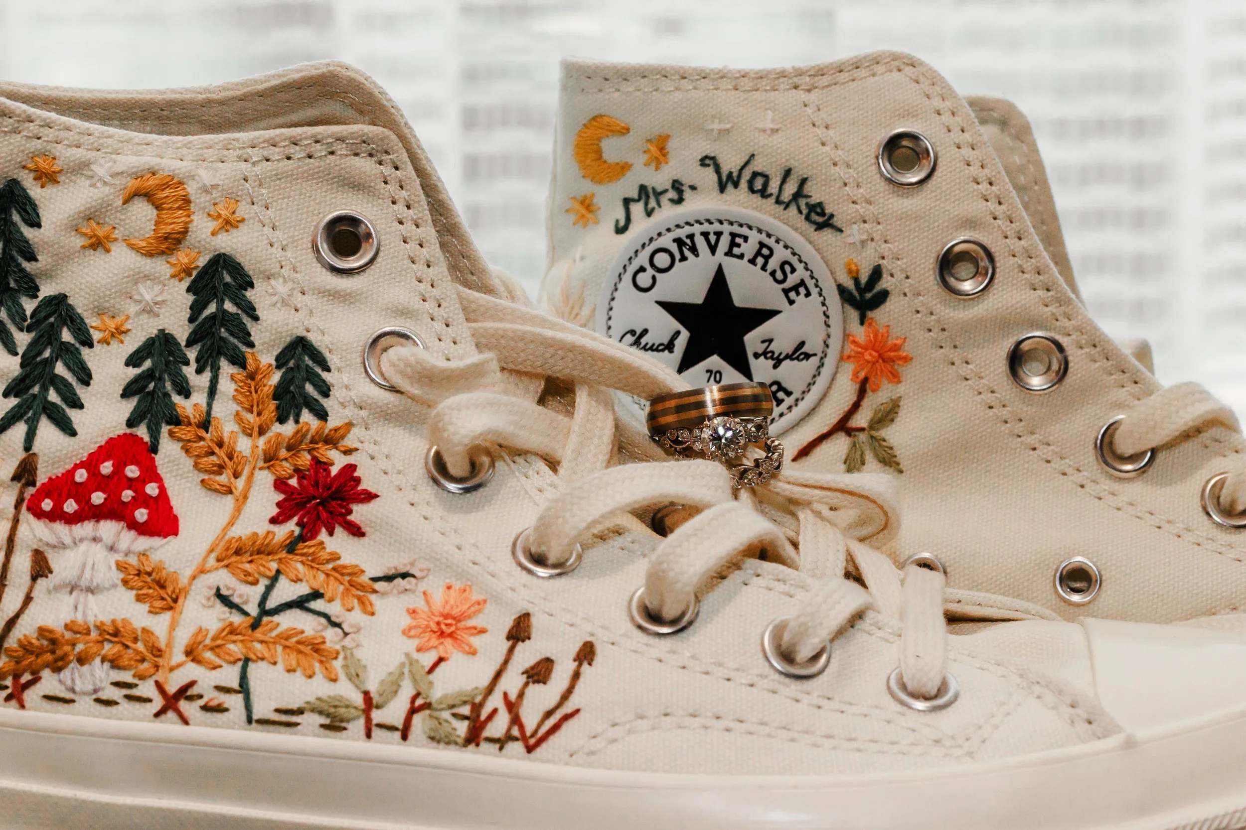 Close-up of embroidered white Converse sneakers with floral designs, featuring a wedding ring and engagement ring placed on the laces.