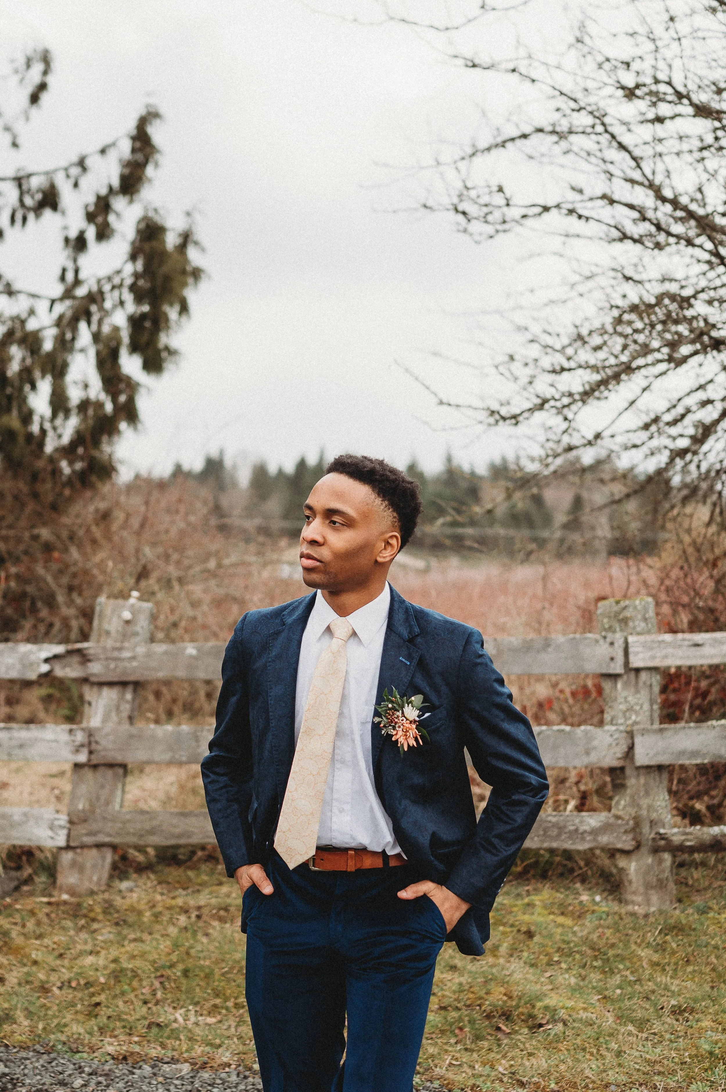 A black man in a blue suit with a boutonniere, standing outdoors in front of a wooden fence and bare trees, looking to the side.