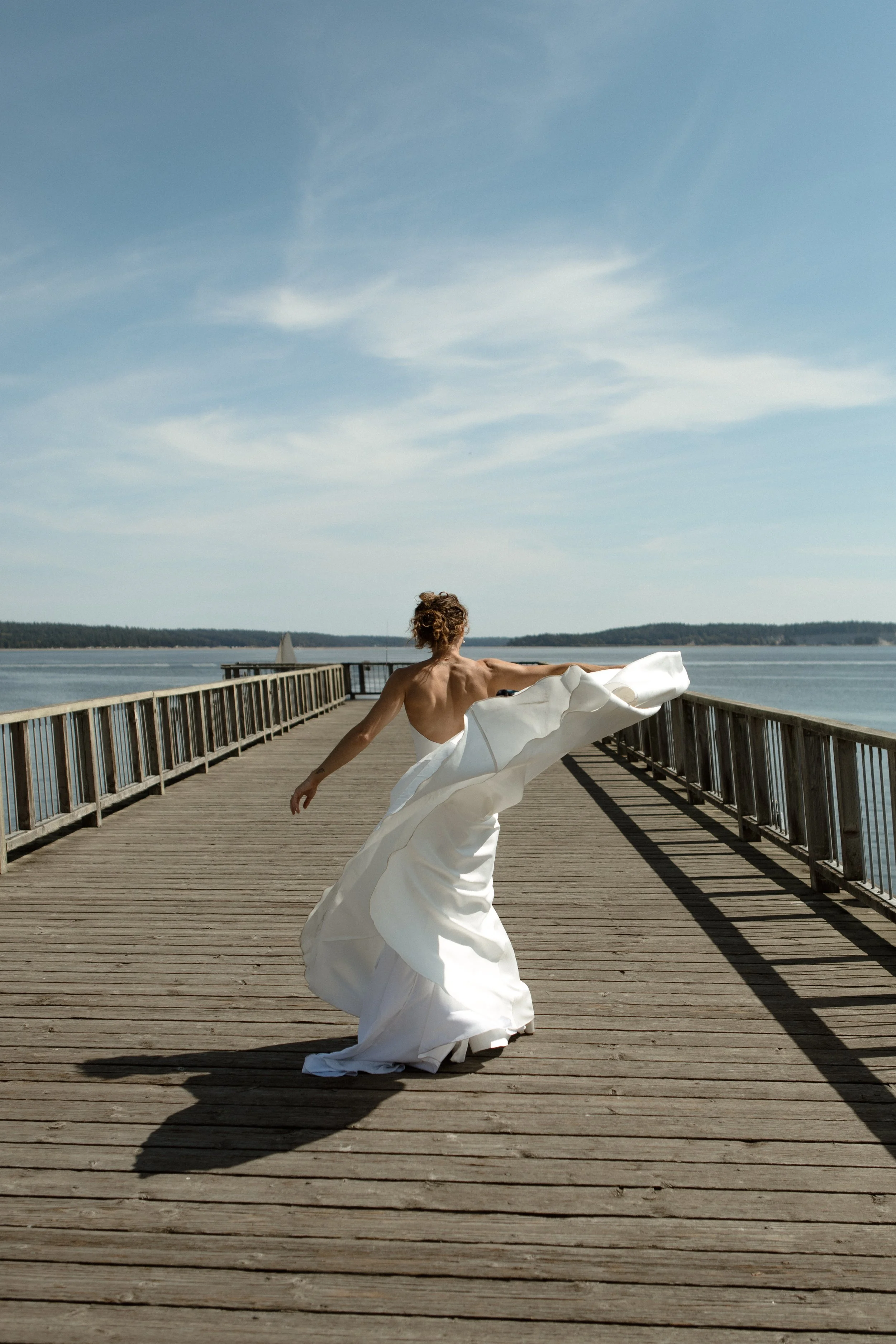 A woman in a white gown spinning on a wooden pier over water, with a blue sky and distant land in the background.