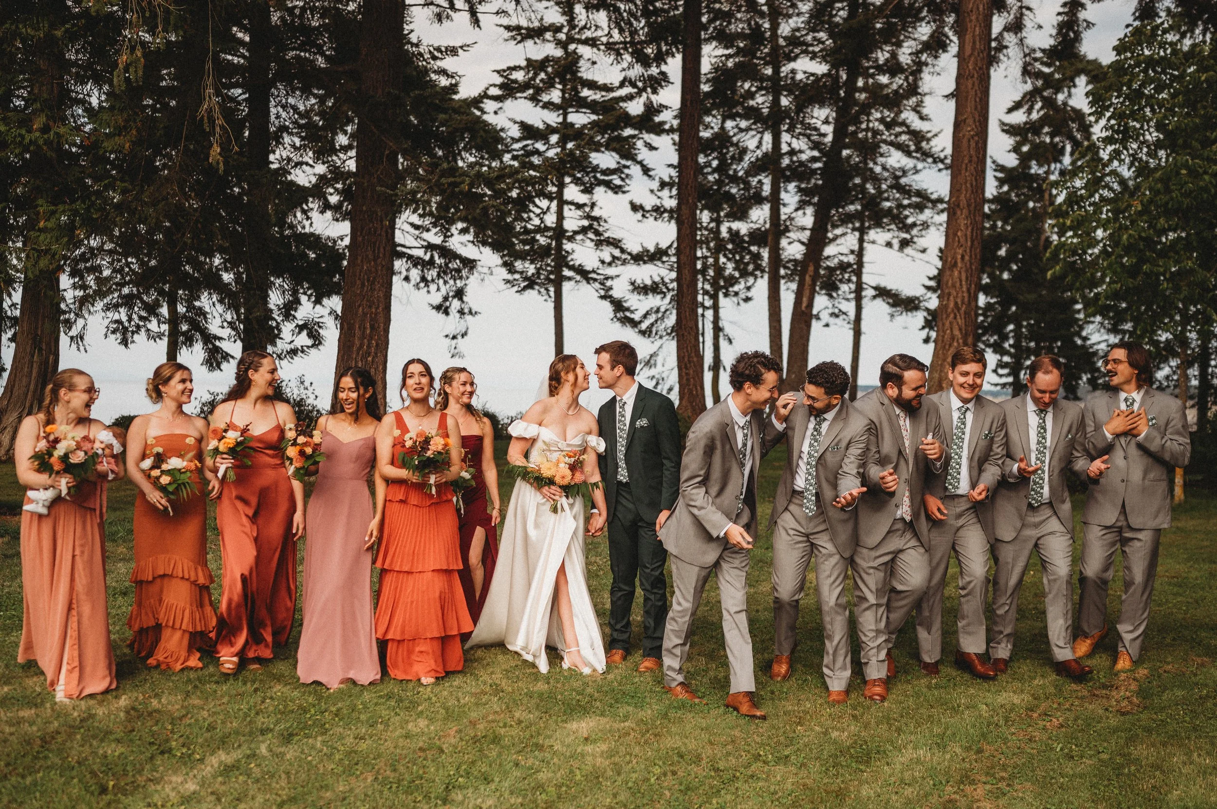 A wedding party of bridesmaids and groomsmen standing and walking outdoors on grass with tall trees in the background. The bridesmaids are wearing orange and pink dresses, holding bouquets of flowers. The groom and groomsmen are dressed in gray suits with patterned ties, some are laughing and chatting.