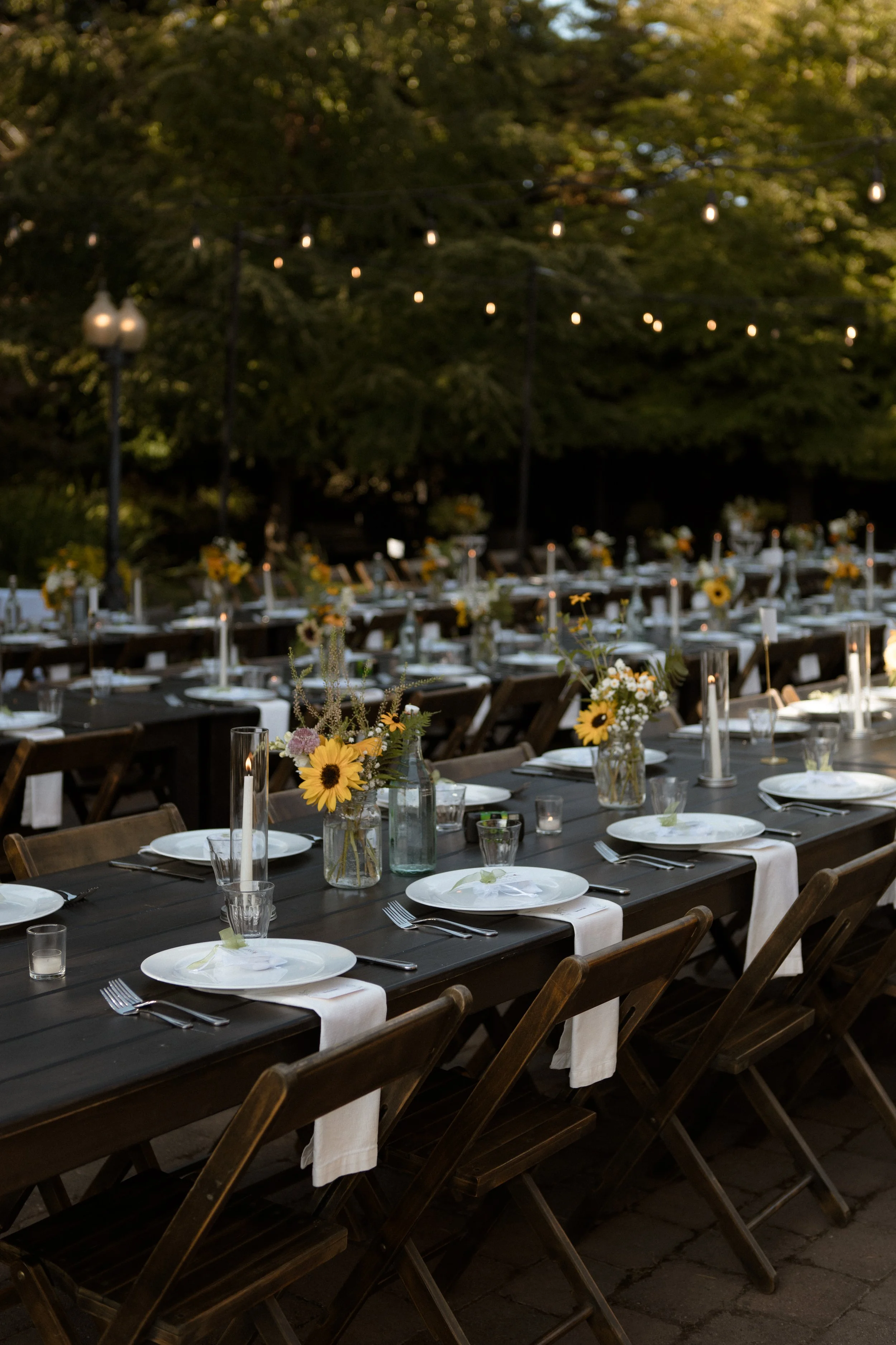Outdoor dining table set for a wedding with white plates, silverware, glassware, and floral centerpieces with sunflowers, under string lights in a garden at dusk.