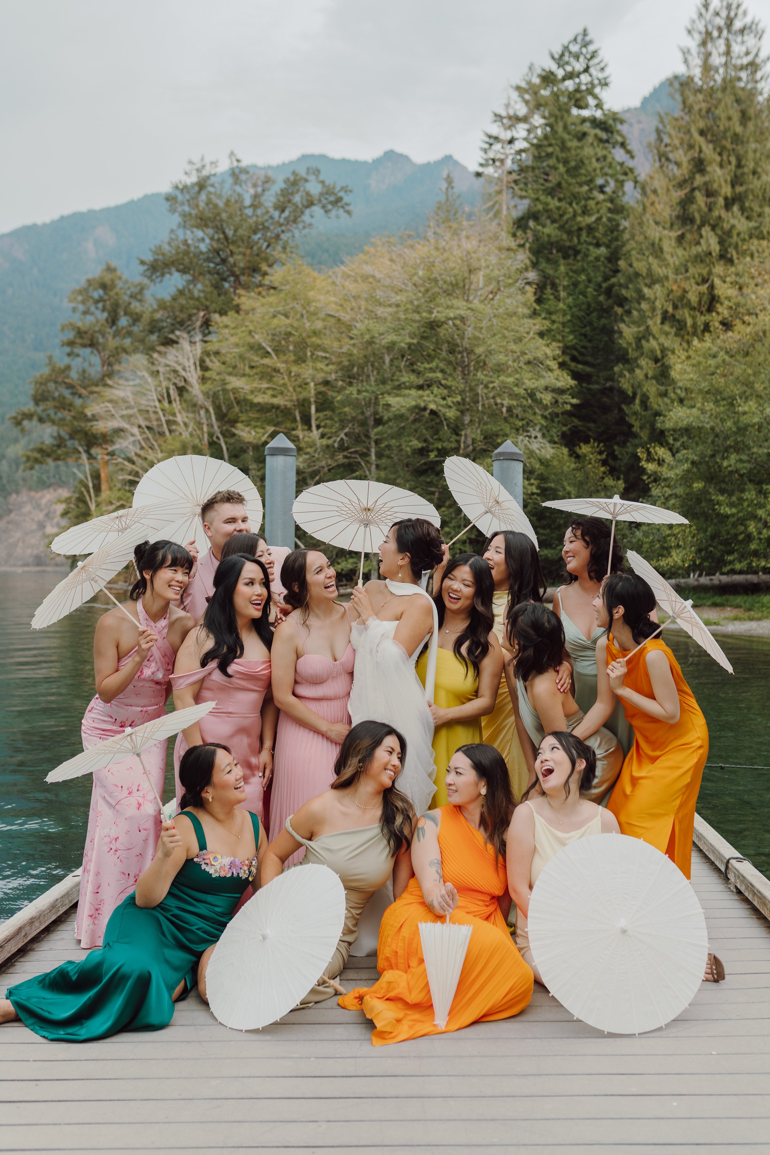 A group of women and one man in colorful dresses holding white parasols, standing on a wooden dock near a lake with forested mountains in the background, celebrating and smiling.