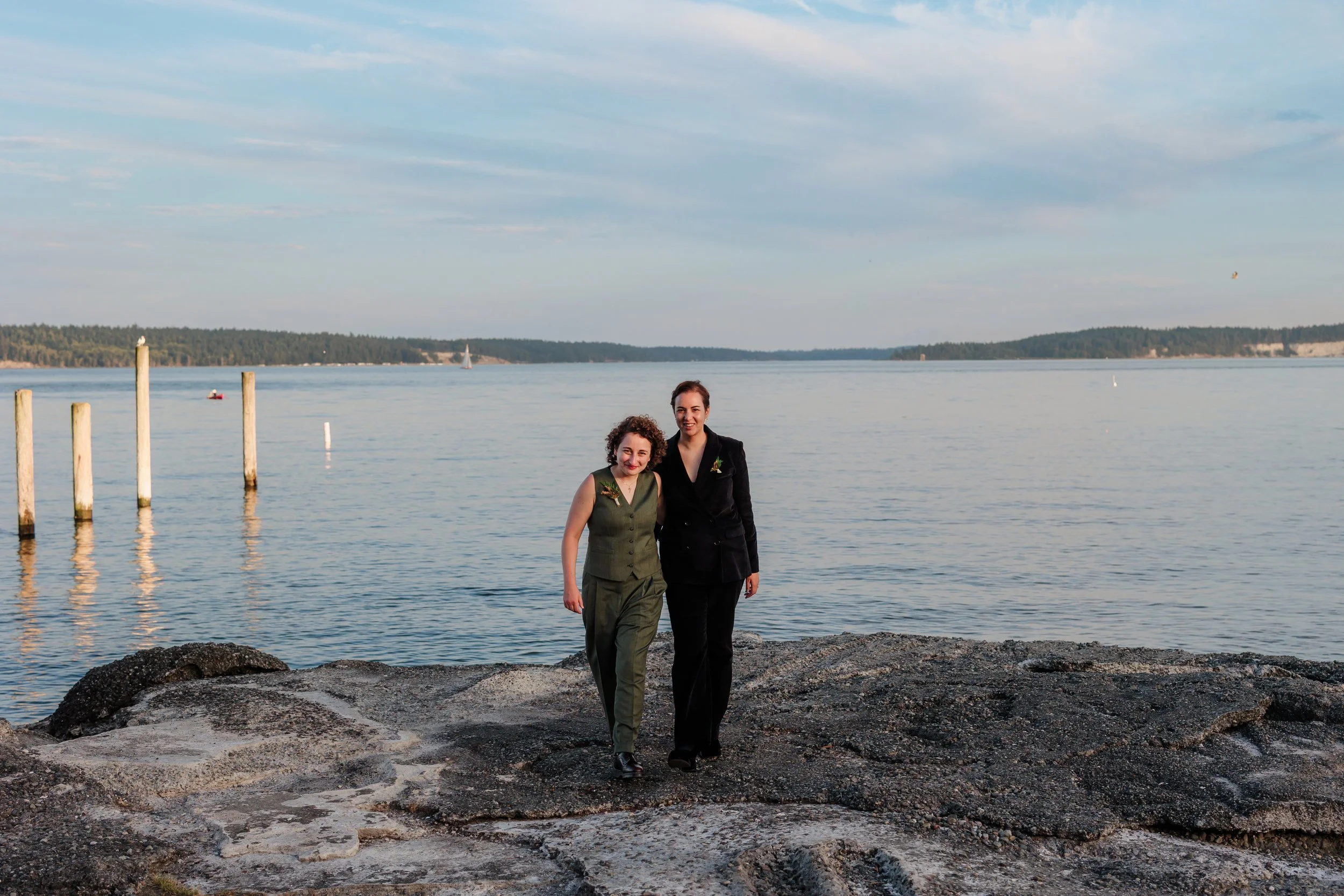 Two women standing on rocky shore on their wedding day, with a distant tree-lined shoreline and sailboats in the background, under a partly cloudy sky.