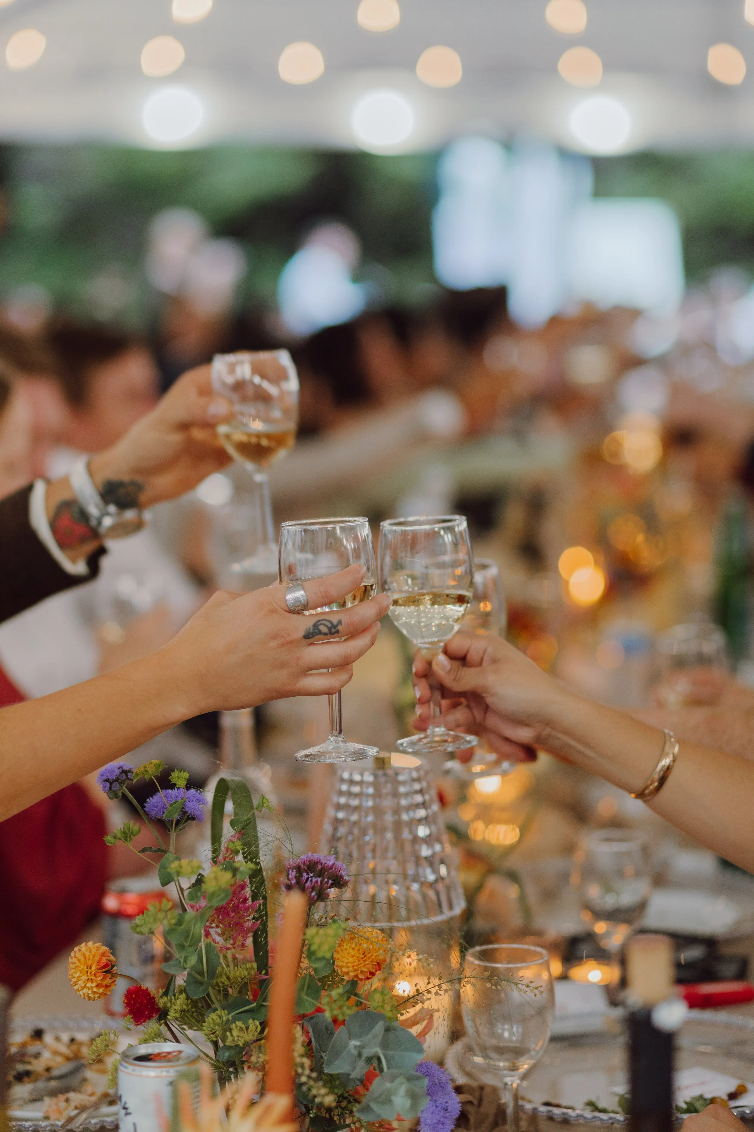 People toasting glasses filled with white wine or champagne at a celebration or banquet table decorated with colorful flowers, candles, and tableware in a well-lit indoor space.