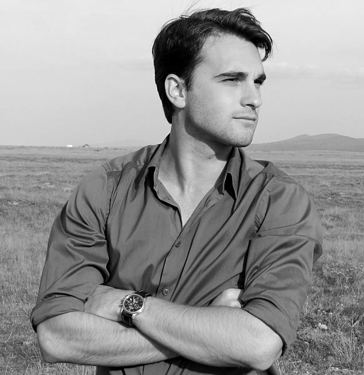 Black and white photo of a young man with dark hair, standing in an open field with mountains in the background, looking to the right with arms crossed, wearing a button-down shirt and a wristwatch.