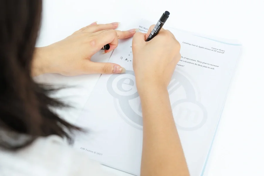 Person writing on a piece of paper with a black marker, with another hand holding the paper steady, on a white surface.