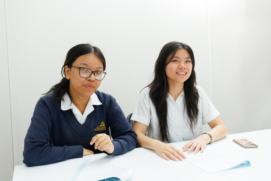 Two smiling students sitting at a white table, one wearing glasses and a navy sweater with a logo, the other in a white shirt, with papers and a smartphone on the table.