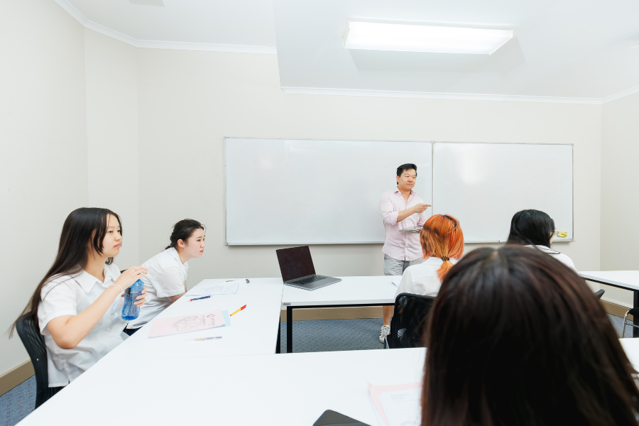 A classroom with students sitting at desks and a teacher standing at the front near a whiteboard.