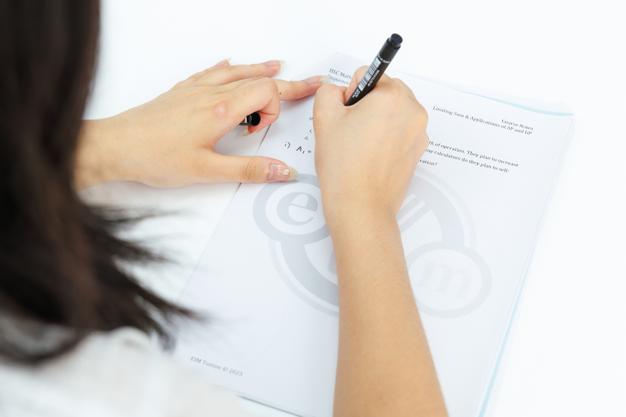 Person writing on a piece of paper with a black marker, with another hand holding the paper steady, on a white surface.