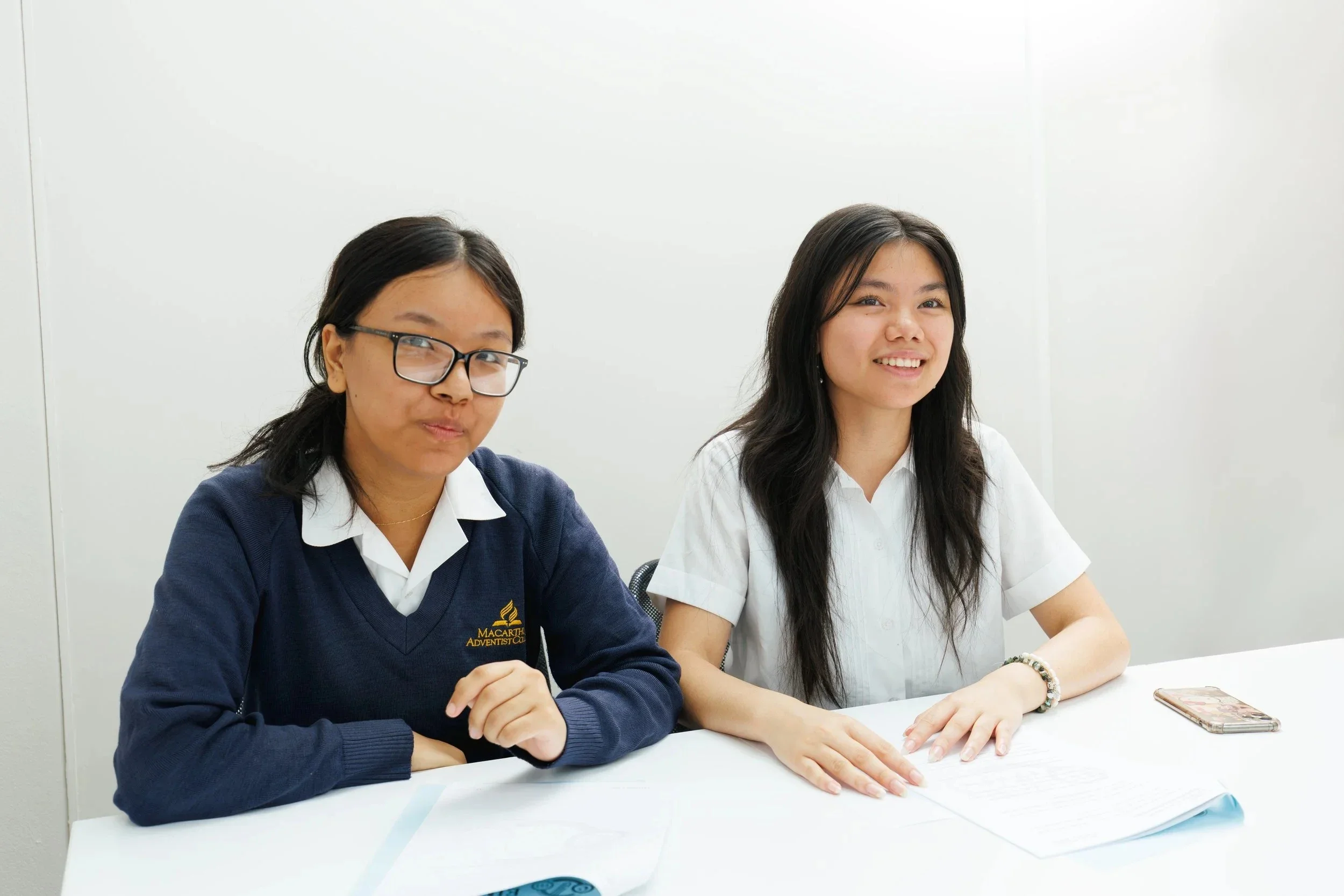 Two smiling students sitting at a white table, one wearing glasses and a navy sweater with a logo, the other in a white shirt, with papers and a smartphone on the table.