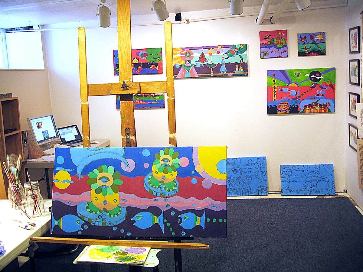 The working studio with finished pieces on the wall, some framed drawings to the right and unfinished canvases on the floor and easel.