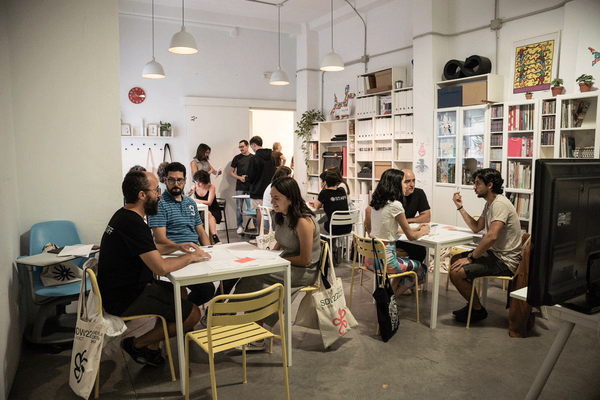 Group of people participating in a discussion or workshop in a modern classroom or community space, with bookshelves, artwork, and white tables.