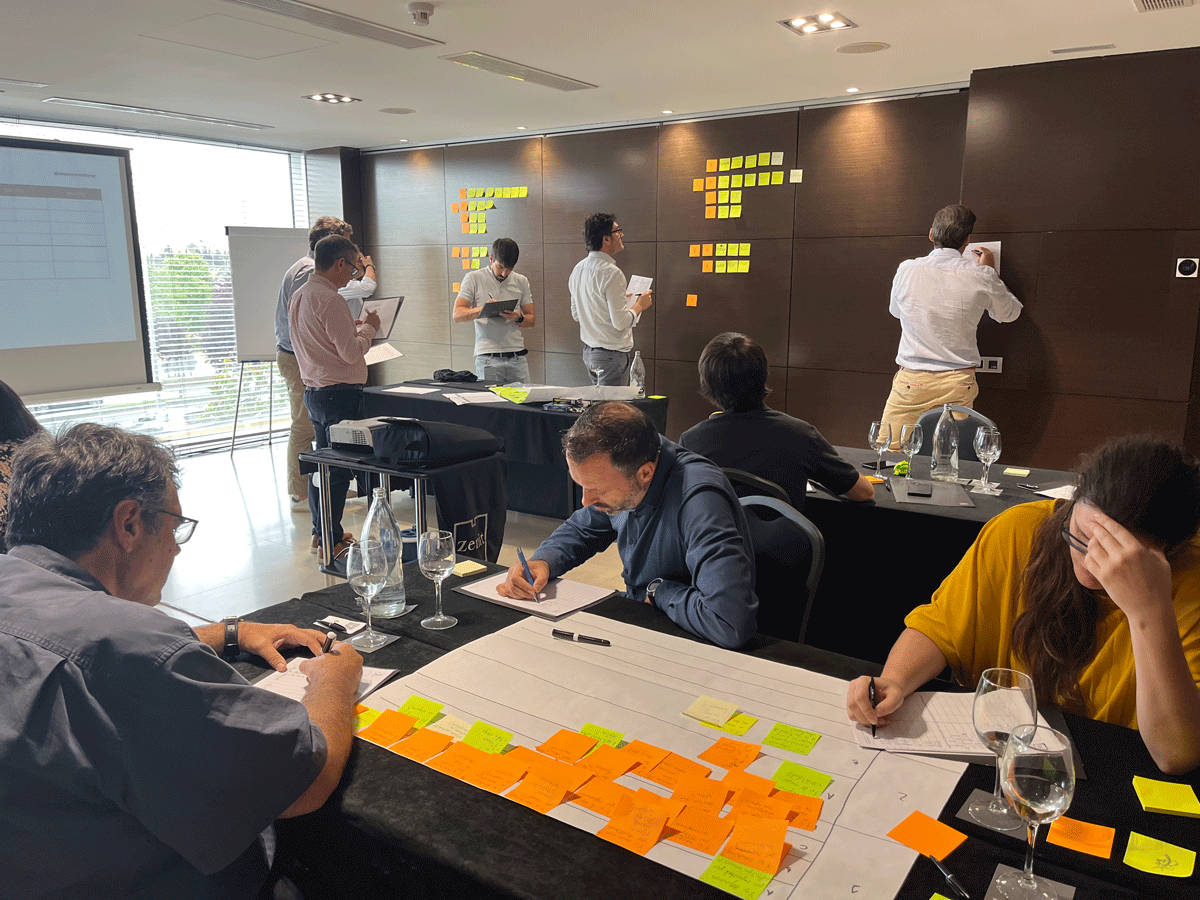 Business meeting in a conference room where people are discussing and writing on sticky notes on a dark wall and tables, with some taking notes and others observing.