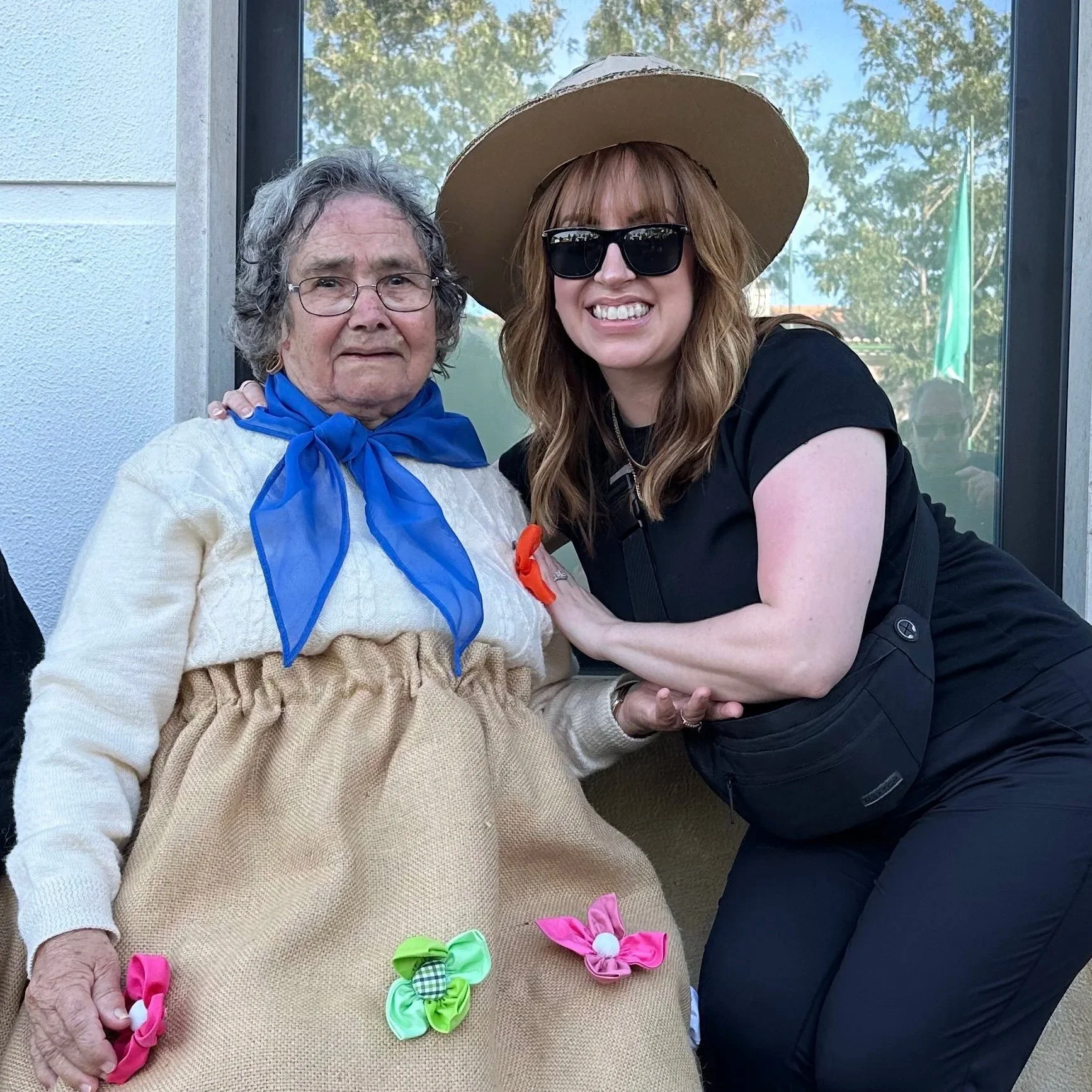 Two women posing together outdoors, one elderly woman wearing a white sweater with a blue scarf, and the other younger woman dressed in black with sunglasses and a wide-brimmed hat. The younger woman is smiling and leaning toward the elderly woman.