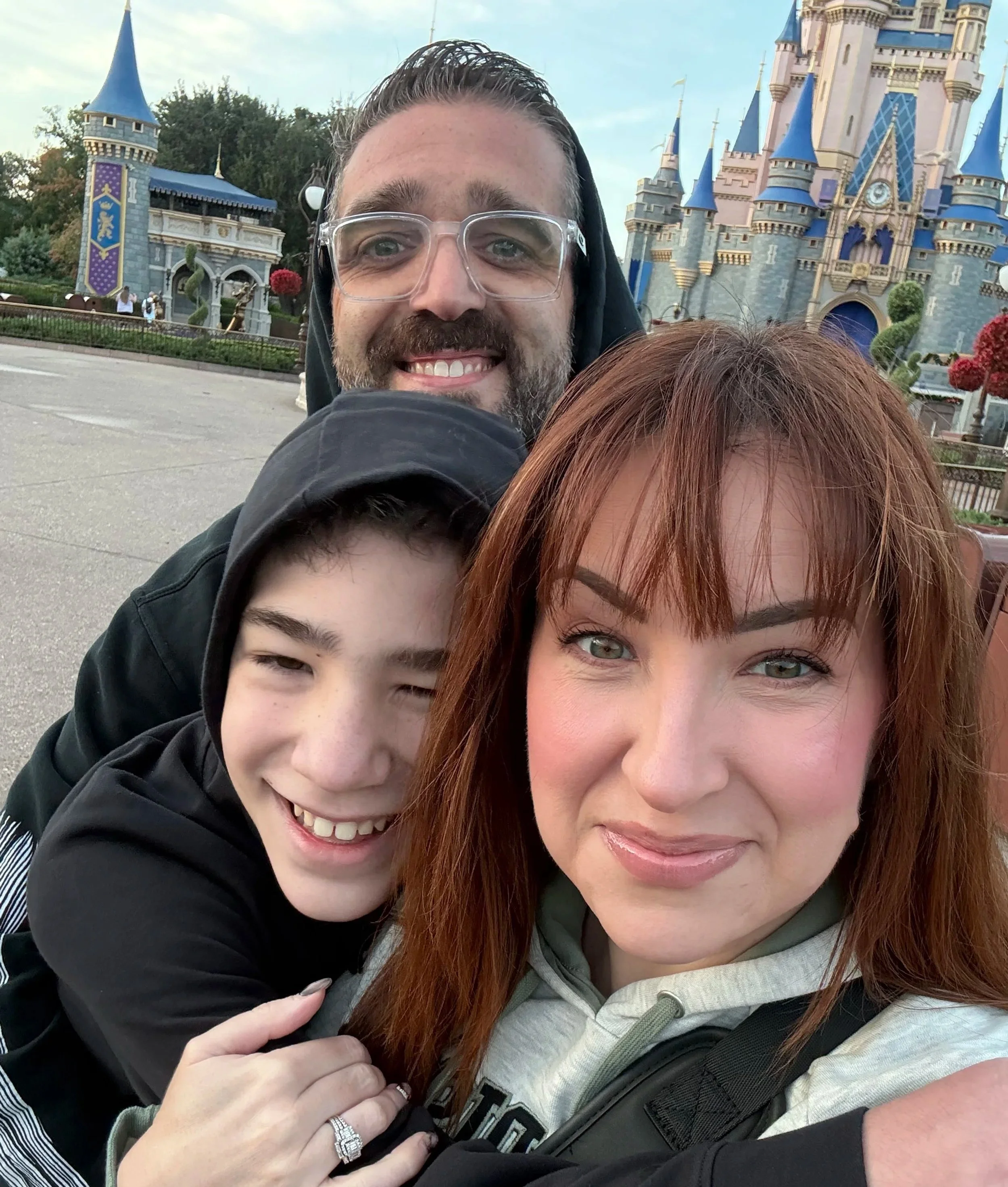 A family of three taking a selfie in front of Disney's castle at Disneyland, with the father smiling behind, the mother with reddish hair in the front, and a young boy with dark hair and a black hoodie.