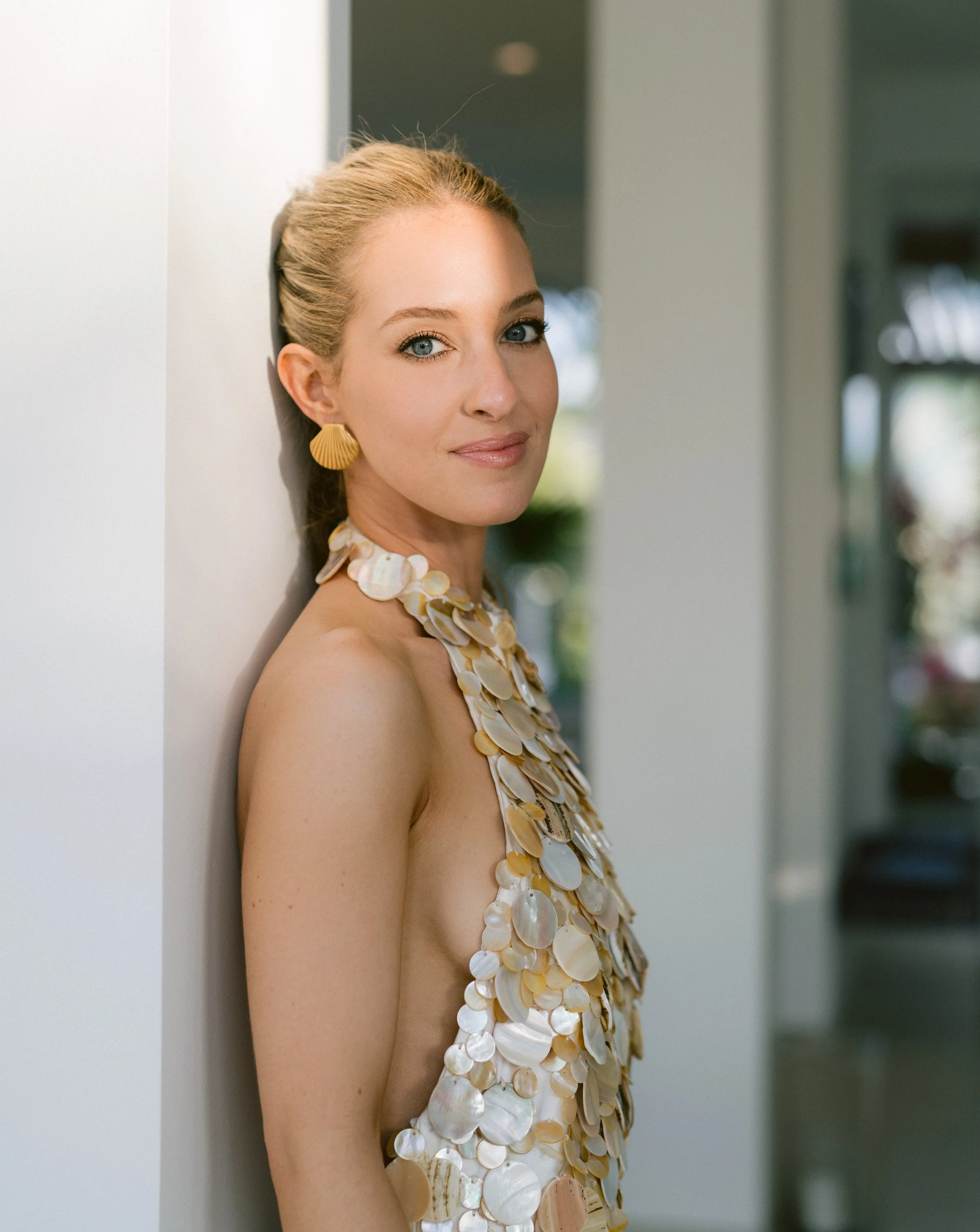 Woman with blonde hair tied back wearing shell jewelry and a shell-covered top, standing against a wall.