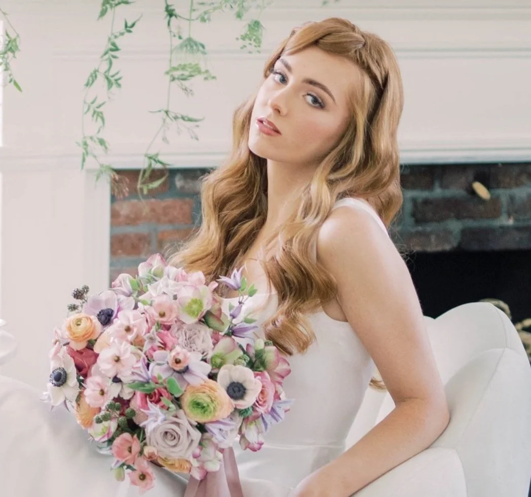 A young woman with long wavy red hair holding a bouquet of pink and purple flowers, sitting on a white chair in front of a white wall with a brick fireplace.