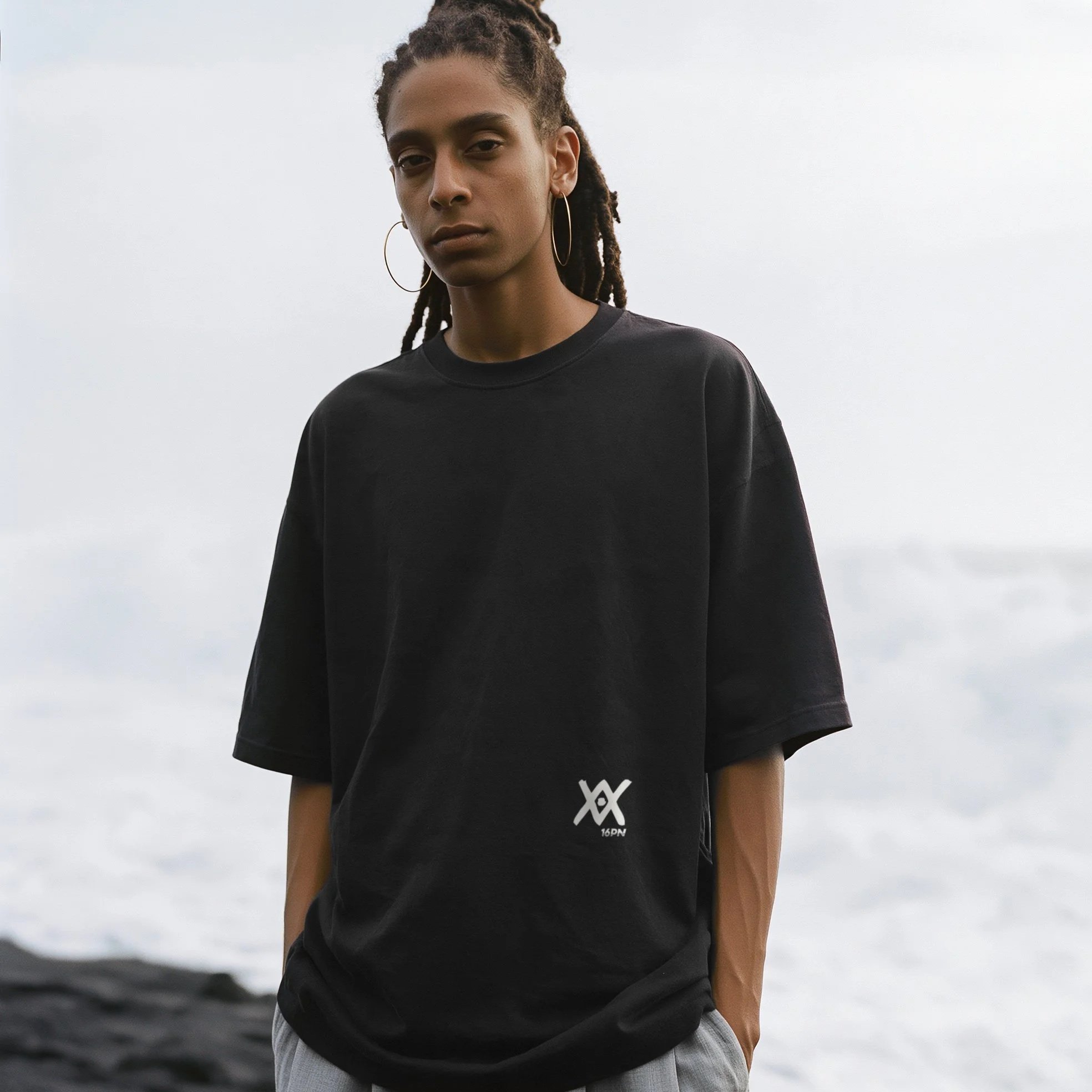 Woman with locs wearing the black 16PN XX oversized tee on a black sand beach in Guadeloupe