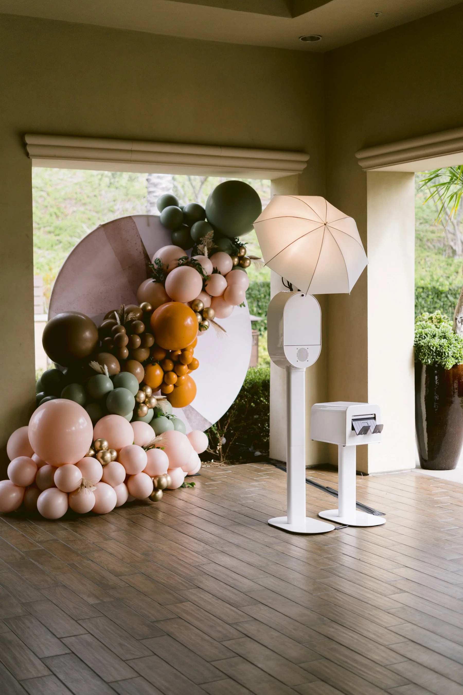 Decorative balloon arrangement with pink, green, gold, and brown balloons, a white umbrella, and a small white table and speaker, set near an open patio with potted plants and outdoor greenery.